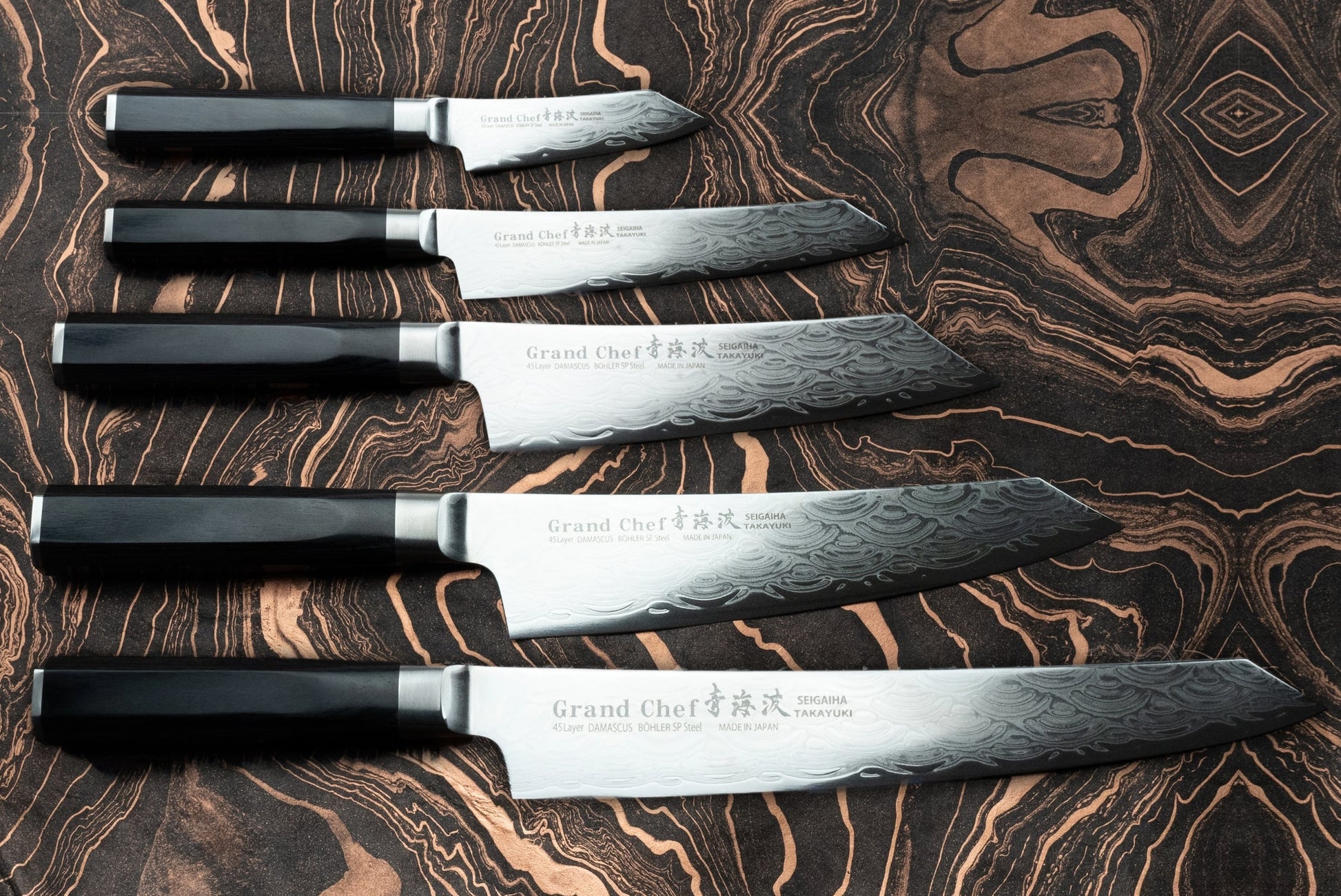 Sakai Takayuki Grand Chef Seigaiha 5 Piece Knife Set displayed with dark handles and patterned Damascus-steel blades, showcasing the “Blue Wave damascus” design on a marbled surface, embodying expert Japanese craftsmanship.