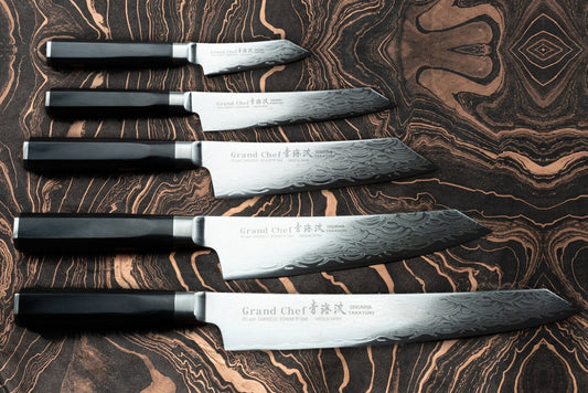 Sakai Takayuki Grand Chef Seigaiha 5 Piece Knife Set displayed with dark handles and patterned Damascus-steel blades, showcasing the “Blue Wave damascus” design on a marbled surface, embodying expert Japanese craftsmanship.