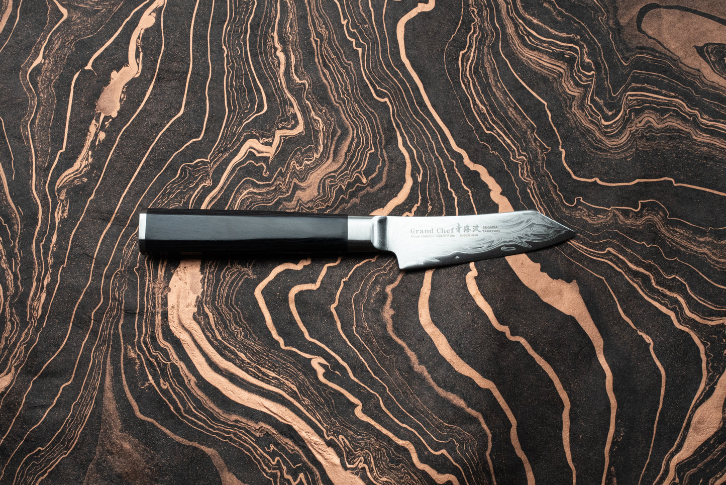 Sakai Takayuki Grand Chef Seigaiha Kengata Petty 80mm knife with an ebony octagonal handle and Damascus-patterned blade, displayed on a marbled surface. Branding and Japanese characters visible on the blade's ricasso.