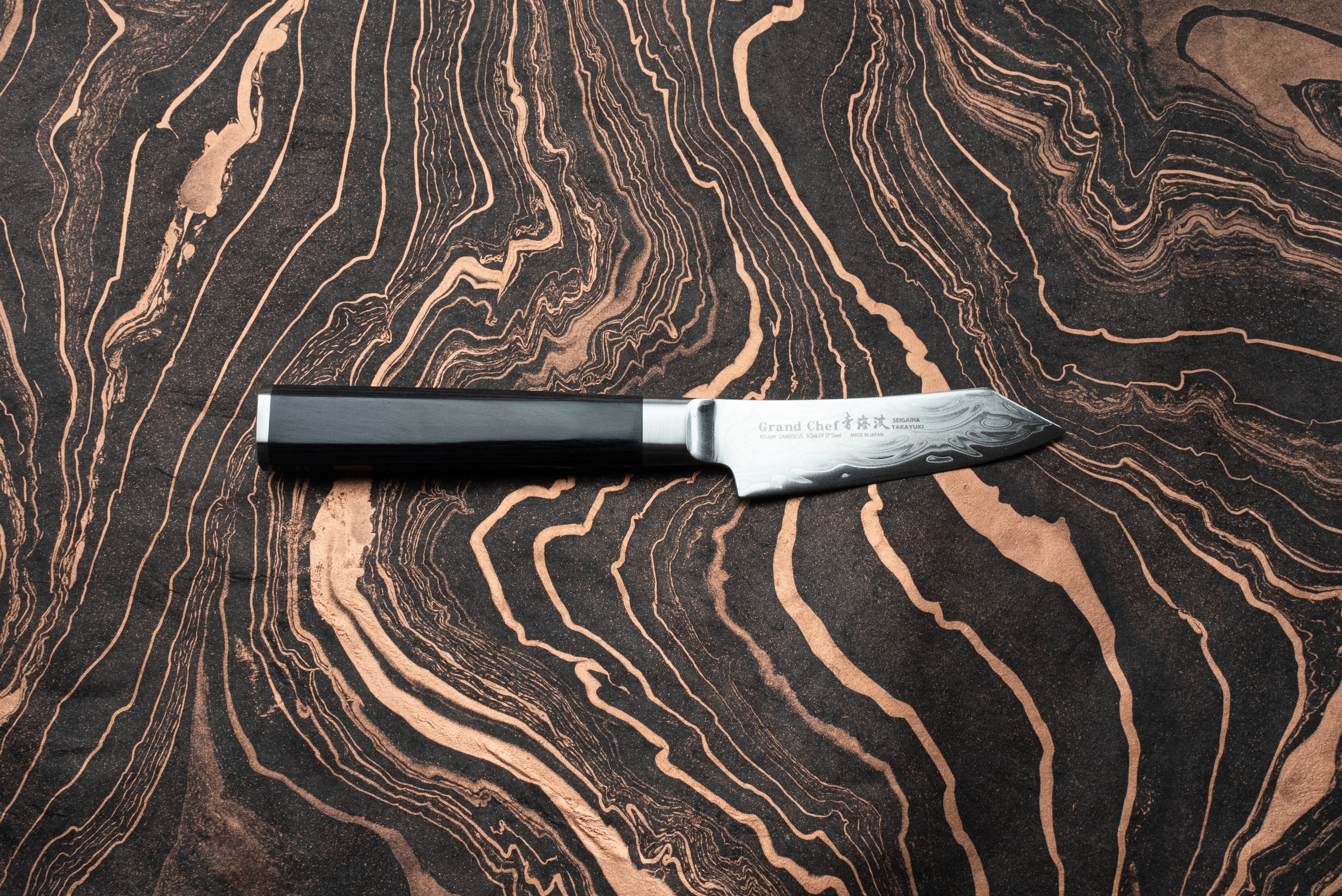 Sakai Takayuki Grand Chef Seigaiha Kengata Petty 80mm knife with an ebony octagonal handle and Damascus-patterned blade, displayed on a marbled surface. Branding and Japanese characters visible on the blade's ricasso.