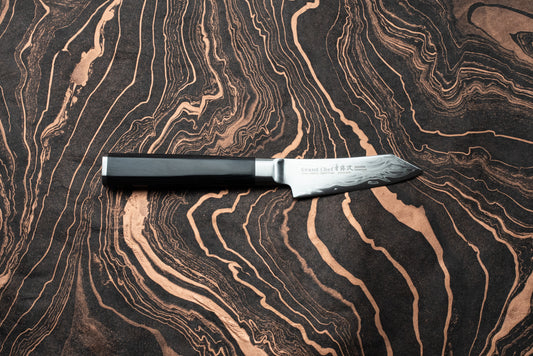 Sakai Takayuki Grand Chef Seigaiha Kengata Petty 80mm knife with an ebony octagonal handle and Damascus-patterned blade, displayed on a marbled surface. Branding and Japanese characters visible on the blade's ricasso.