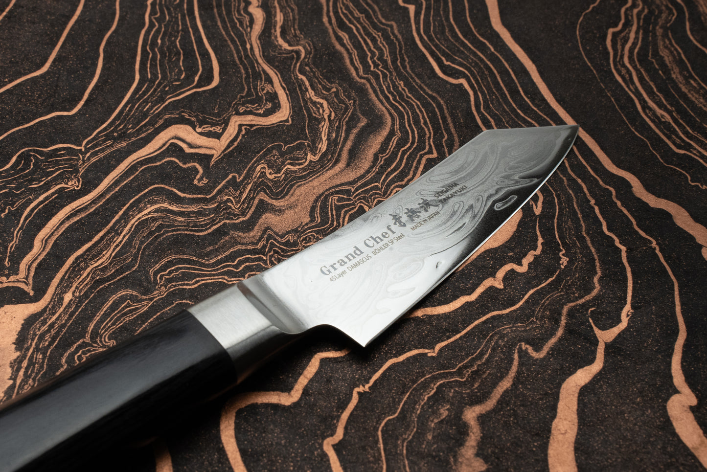 Sakai Takayuki Grand Chef Seigaiha Kengata Petty 80mm knife on a marbled surface, showcasing its Damascus-patterned steel blade etched with Grand Chef and Japanese characters, featuring a dark wood handle.