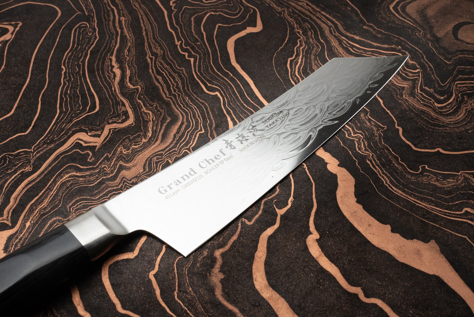 Sakai Takayuki Grand Chef Seigaiha Kengata Santoku 160mm knife blade, showcasing a 45-layer damascus pattern, lies on a wood-grain surface, with a partially visible handle and prominent etched design.