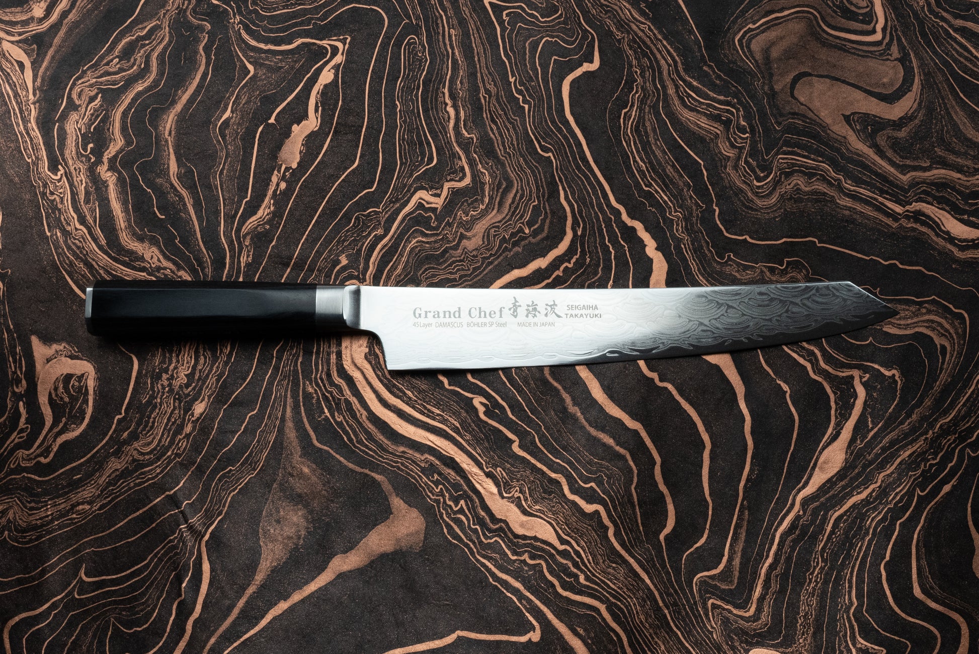 Sakai Takayuki Grand Chef Seigaiha Kengata Sujihiki 250mm knife with a Damascus-patterned blade and an etched logo, featuring a sleek, dark octagonal handle, displayed on a marbled swirl-patterned surface.