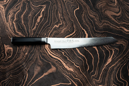 Sakai Takayuki Grand Chef Seigaiha Kengata Sujihiki 250mm knife with a Damascus-patterned blade and an etched logo, featuring a sleek, dark octagonal handle, displayed on a marbled swirl-patterned surface.
