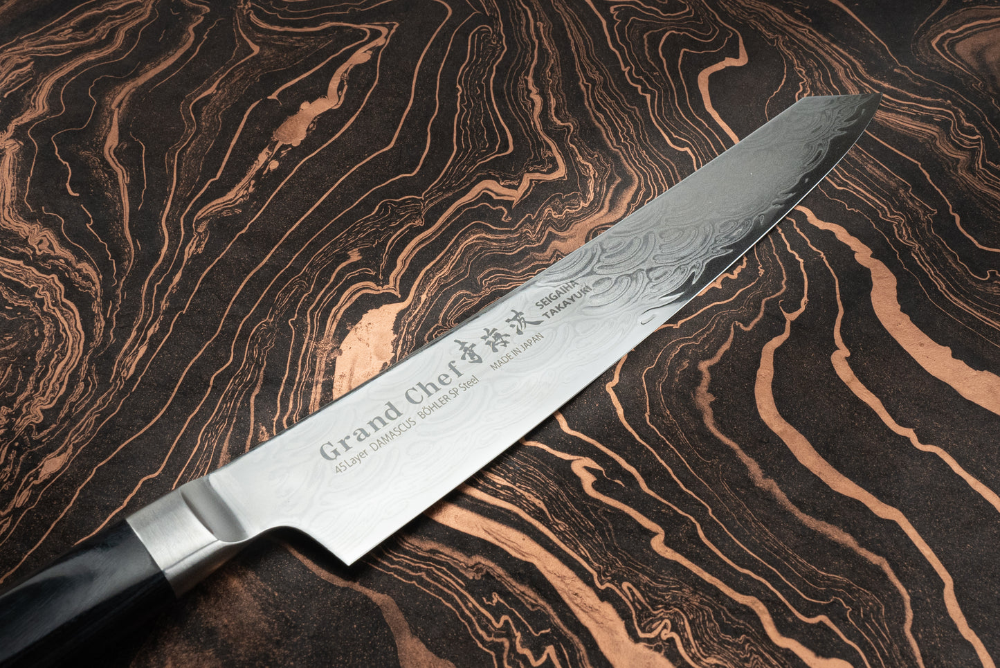 Sakai Takayuki Grand Chef Seigaiha Kengata Sujihiki 250mm knife with 45-layer Damascus Böhler SP steel blade and polished hardwood handle, showcasing intricate etched patterns and craftsmanship, laid on a marbled background.