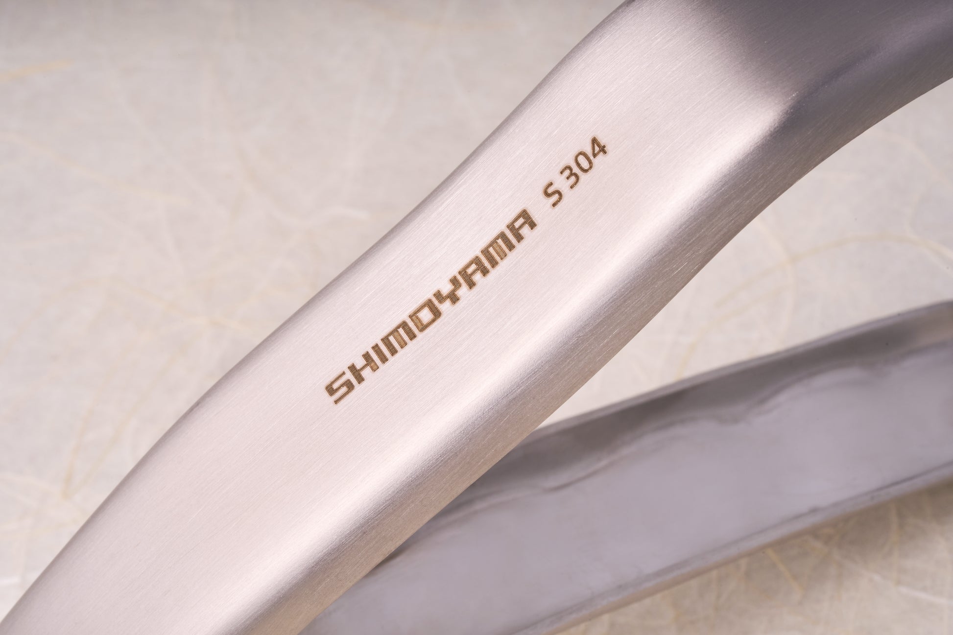 Close-up of Shimoyama Stainless Steel Bowl Clip, showcasing etched SHIMOHAMA S304 on brushed stainless steel, highlighting its fine grain and gentle curve.