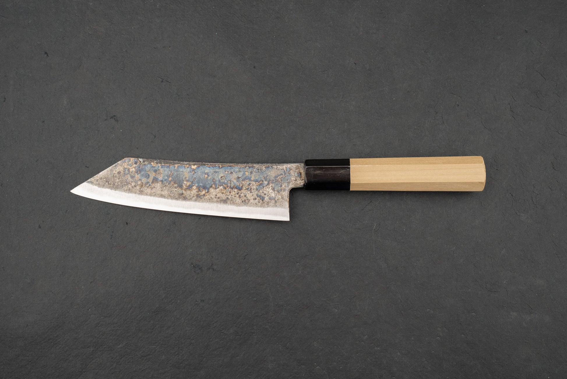 Shiro Kunimitsu Aogami Kurouchi Chikuzen Bunka 160mm knife on slate, showcasing its hammered blade finish and polished edge, with an octagonal light-wood handle and dark collar, embodying traditional craftsmanship.