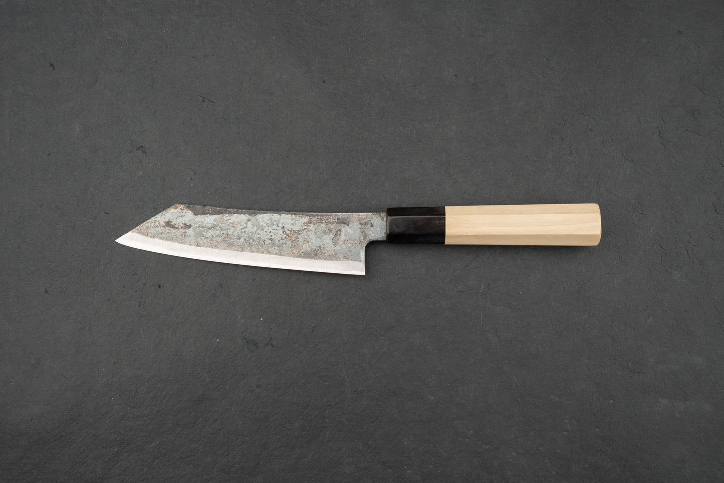 Shiro Kunimitsu Aogami Kurouchi Chikuzen Ko-Bunka 140mm knife with a rustic hammered blade and traditional octagonal wooden handle, displayed on a dark surface, embodying Japanese craftsmanship.
