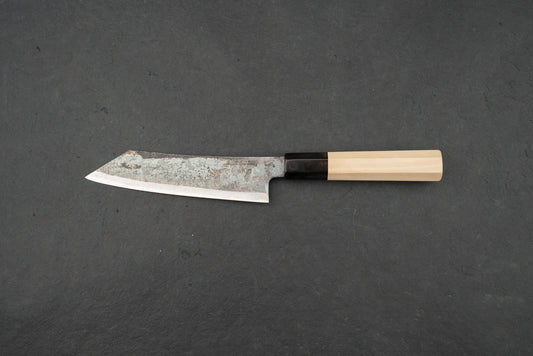 Shiro Kunimitsu Aogami Kurouchi Chikuzen Ko-Bunka 140mm knife with a rustic hammered blade and traditional octagonal wooden handle, displayed on a dark surface, embodying Japanese craftsmanship.