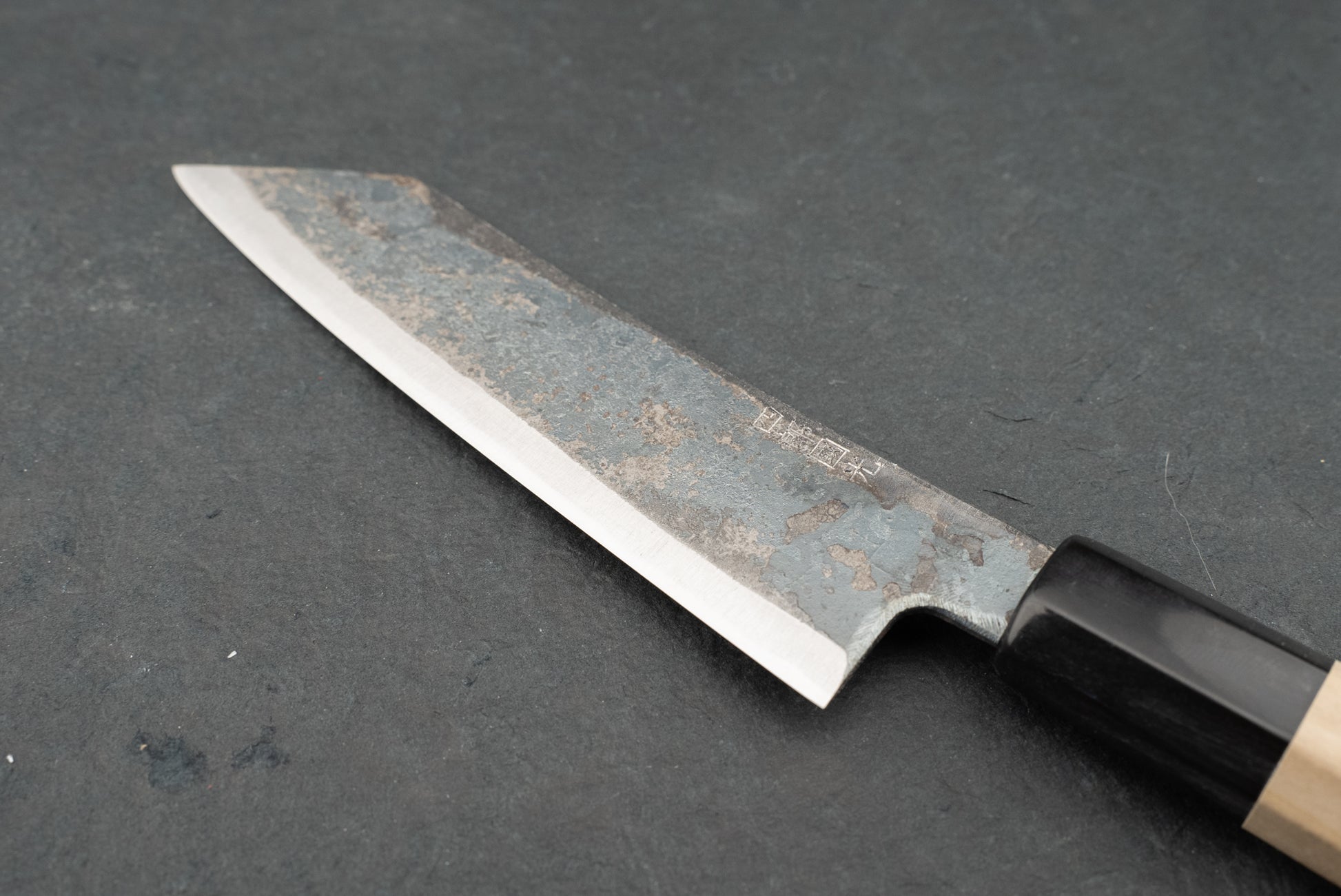 Shiro Kunimitsu Aogami Kurouchi Chikuzen Ko-Bunka 140mm knife on slate, showcasing its single-bevel edge, hammer-textured blade, octagonal wooden handle, and kanji engraving, epitomizing traditional Japanese craftsmanship.