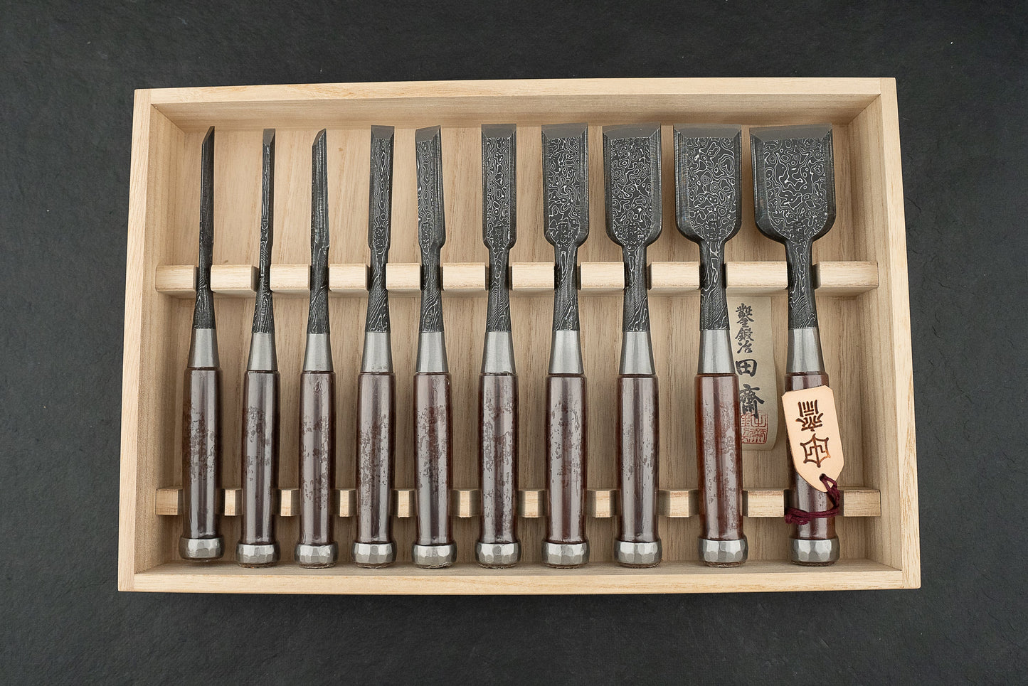 Tasai Aogami Mokume Damascus chisels in a wooden box, featuring ten laminated steel blades with distinctive patterns and turned wooden handles, embody precision craftsmanship for Japanese woodworking enthusiasts.