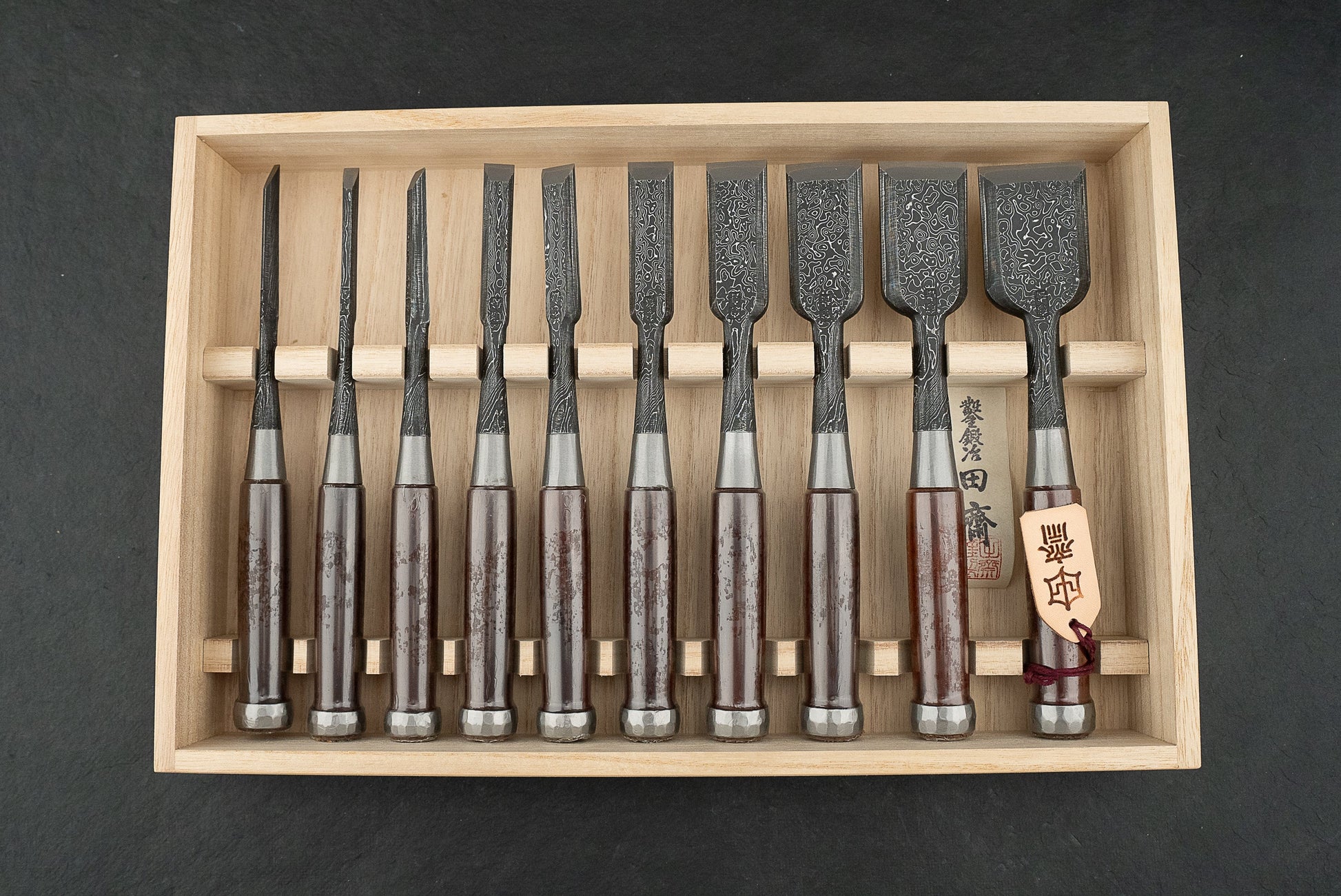 Tasai Aogami Mokume Damascus chisels in a wooden box, featuring ten laminated steel blades with distinctive patterns and turned wooden handles, embody precision craftsmanship for Japanese woodworking enthusiasts.