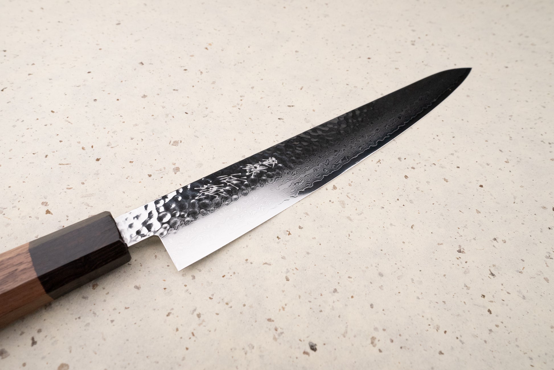Touroku Sakai Yukage Sujihiki 240mm, a Japanese kitchen knife with a hammered Damascus blade and octagonal wooden handle, lies elegantly on a countertop, embodying artisanal craftsmanship for precise meat slicing.