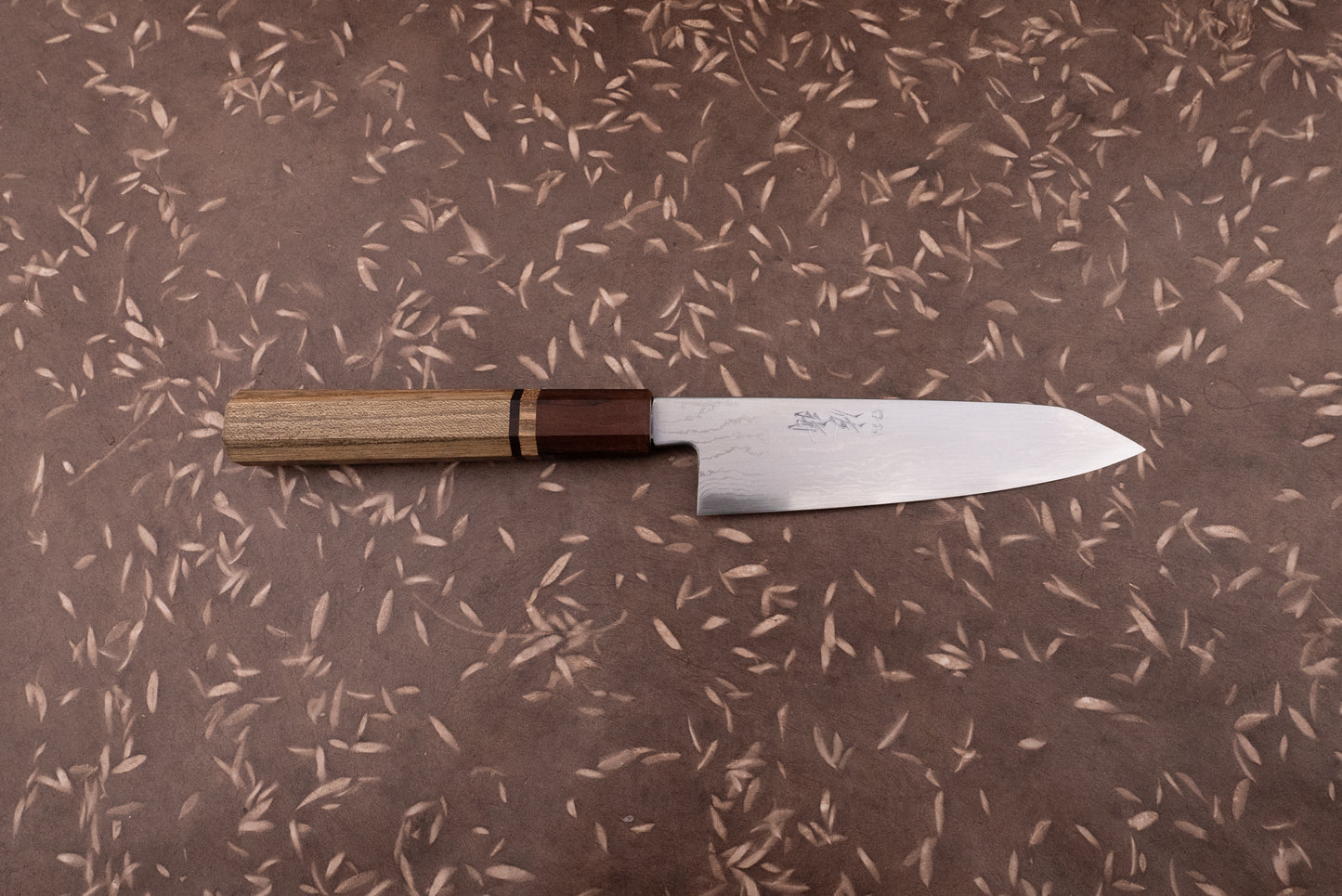 Touroku Sakai Aogami Damascus Kiritsuke Petty 150mm knife with a laminated steel blade and octagonal wooden handle on a brown surface with leaf motifs, ideal for precise kitchen tasks.