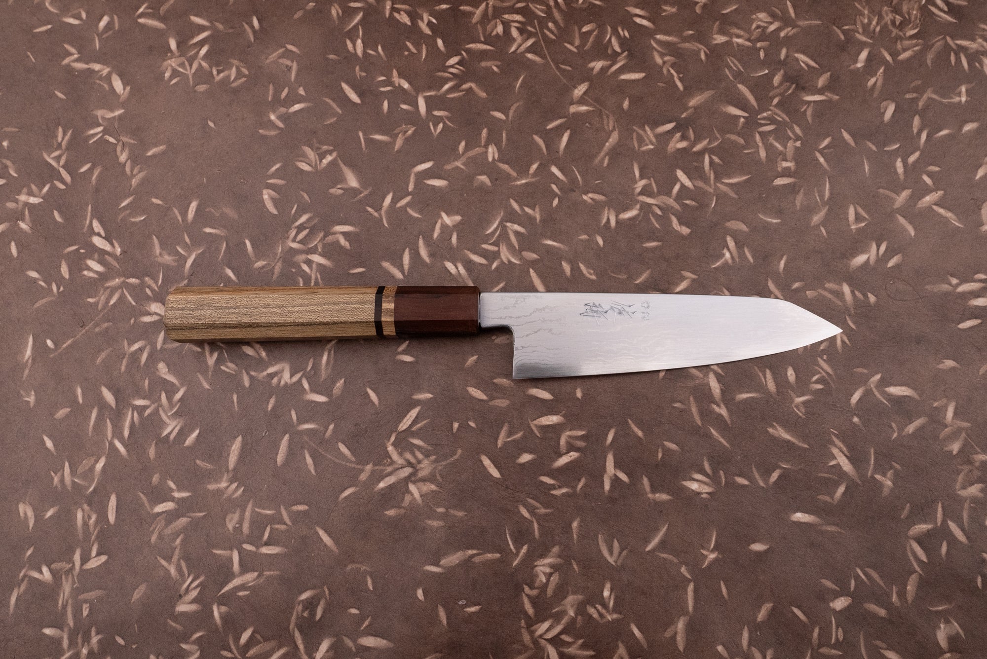 Touroku Sakai Aogami Damascus Kiritsuke Petty 150mm knife with a laminated steel blade and octagonal wooden handle on a brown surface with leaf motifs, ideal for precise kitchen tasks.