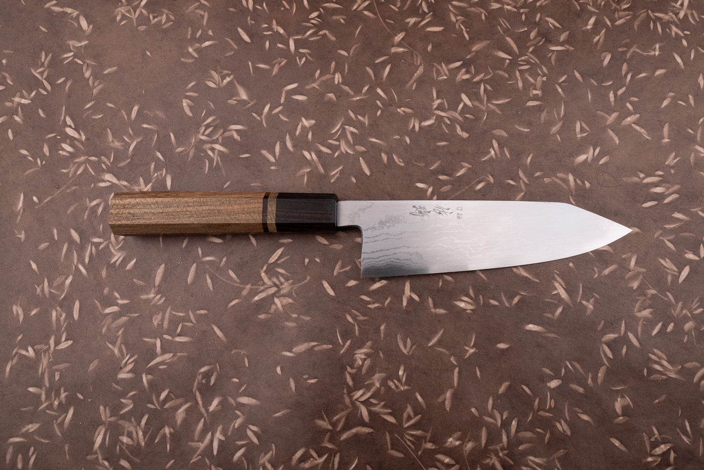 Touroku Sakai Aogami Damascus Kiritsuke Santoku 180mm knife with a laminated steel blade and kanji inscriptions, featuring an octagonal Wa-style handle of contrasting woods, displayed on a textured surface.