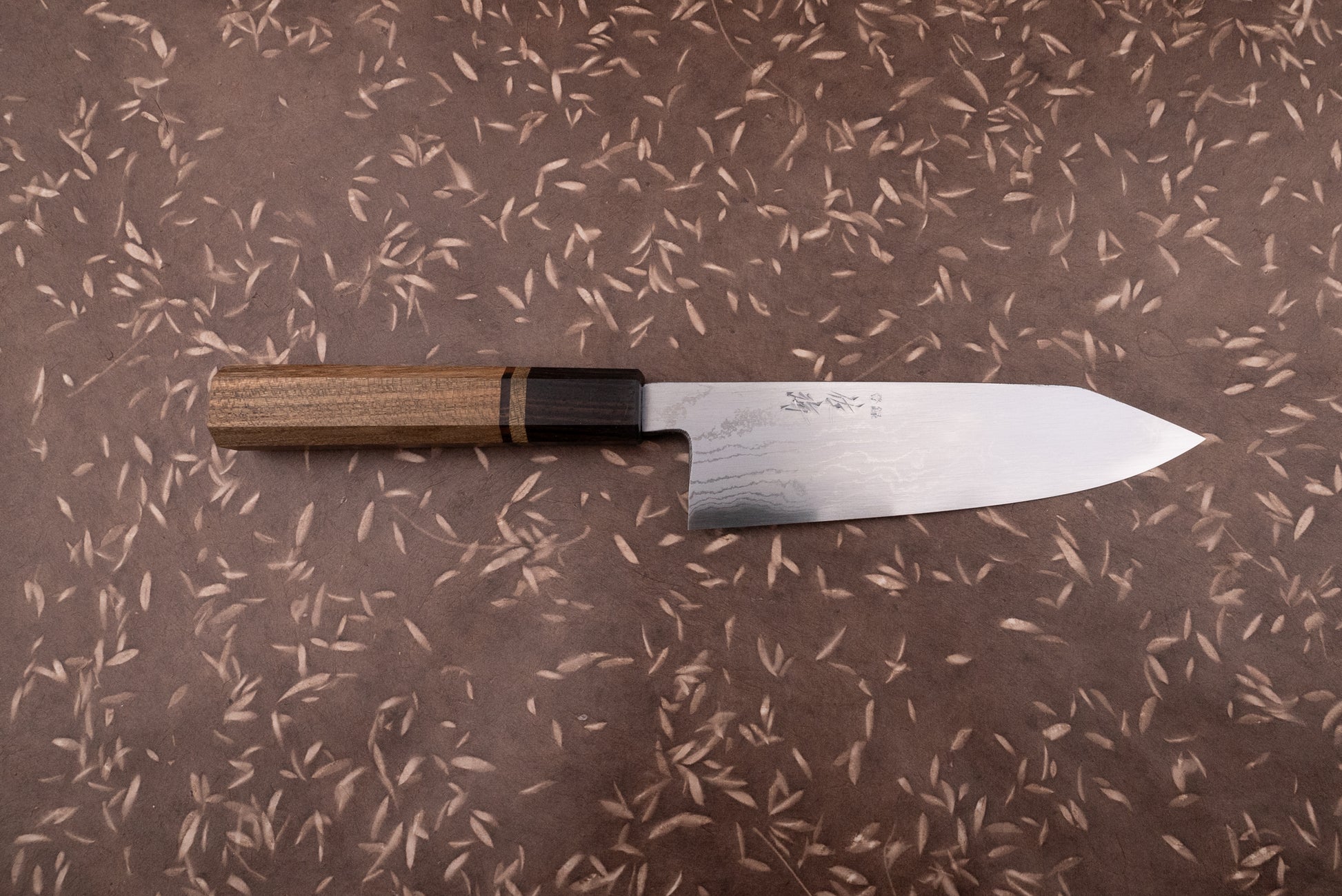 Touroku Sakai Aogami Damascus Kiritsuke Santoku 180mm knife with a laminated steel blade and kanji inscriptions, featuring an octagonal Wa-style handle of contrasting woods, displayed on a textured surface.
