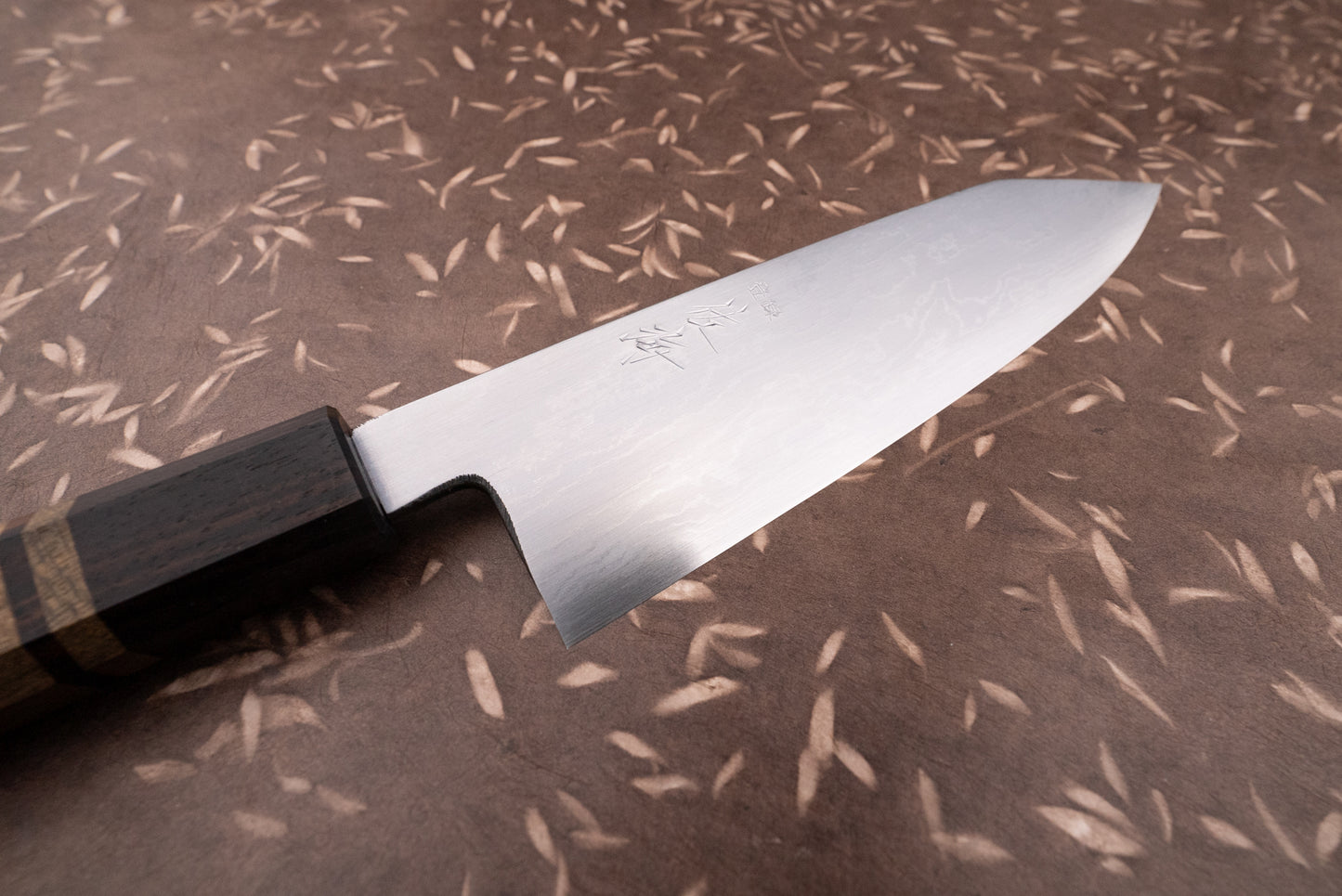 Touroku Sakai Aogami Damascus Kiritsuke Santoku 180mm knife with a polished steel blade featuring damascus grain and engraved Japanese characters, and a dark wooden handle, on a patterned brown surface.