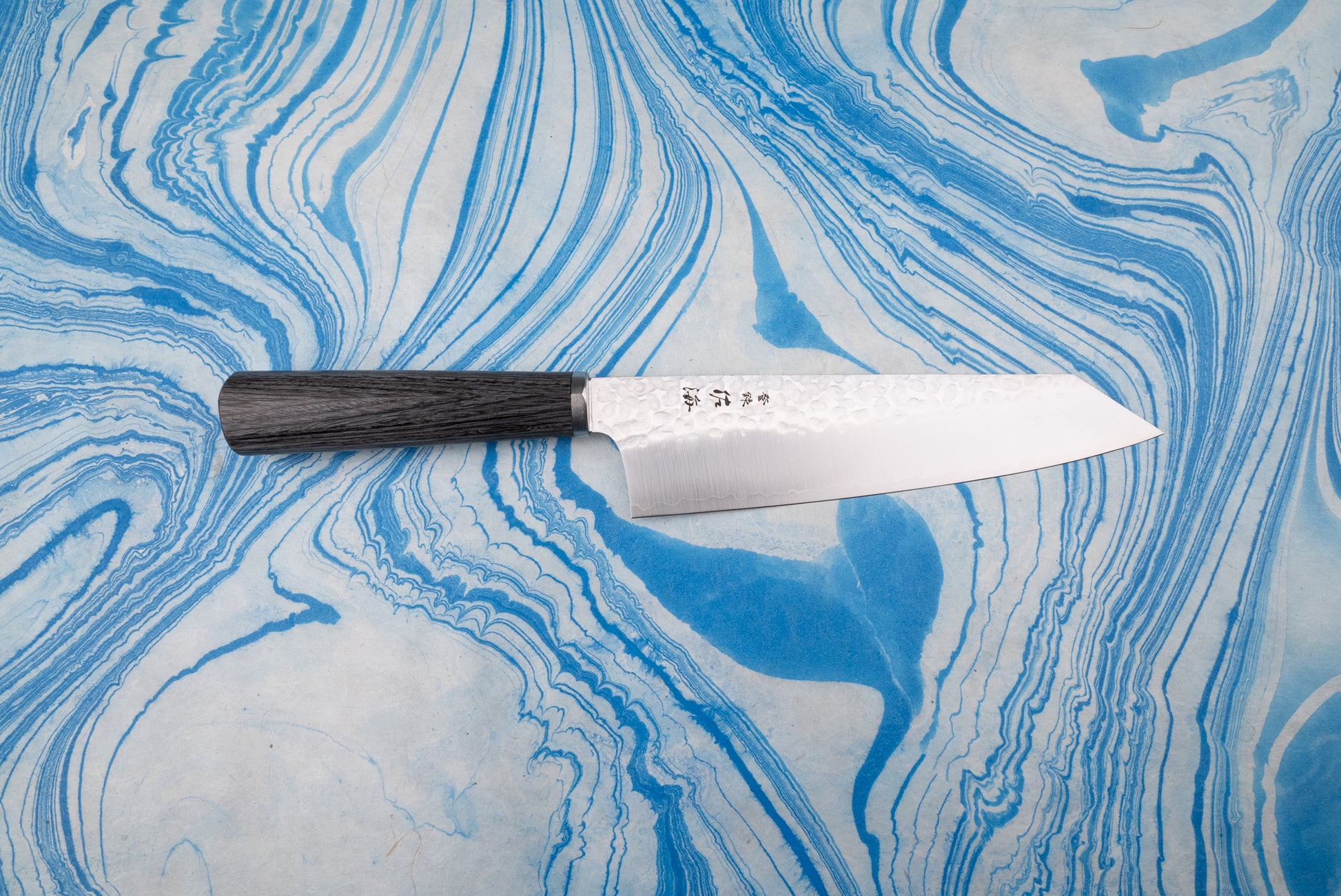 Touroku Sakai Gekka Bunka 180mm, a Japanese kitchen knife with a dark wooden handle and dimpled blade displaying Japanese characters, on a marbled surface, embodies exceptional craftsmanship and balance.