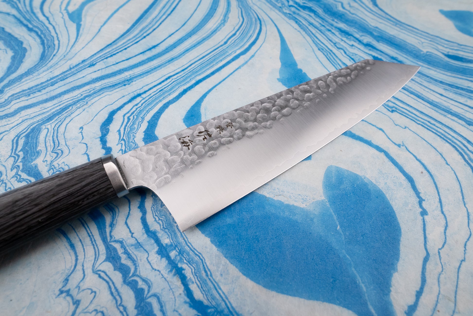 Touroku Sakai Gekka Bunka 180mm knife on a marbled surface, showcasing its hammered steel blade with kanji stamps and a dark wooden handle, embodying Japanese craftsmanship in a versatile kitchen tool.