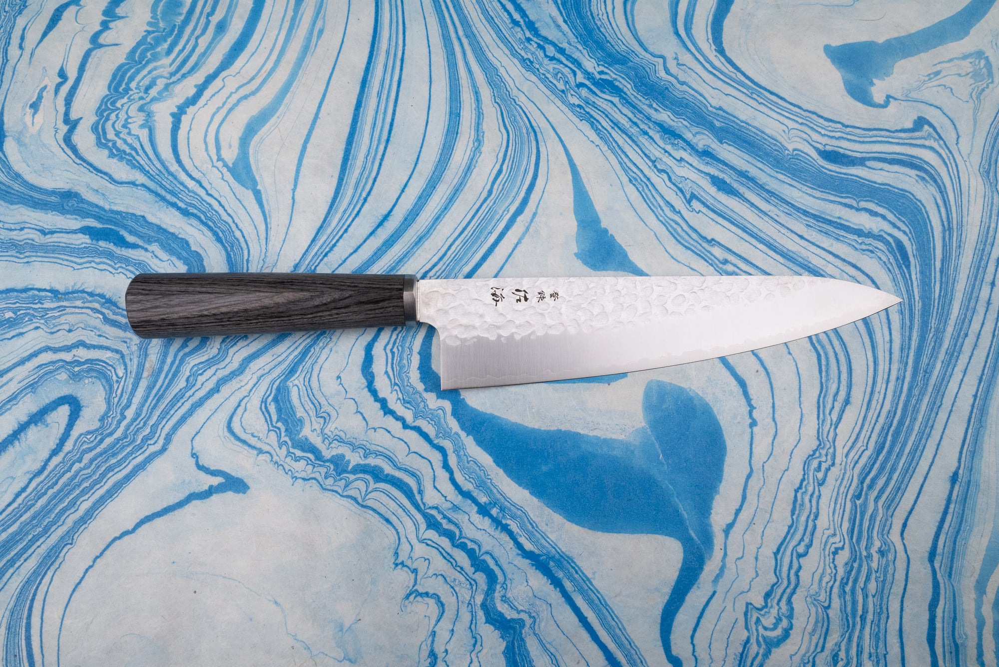 Touroku Sakai Gekka Gyuto 200mm features a hammer-textured stainless-steel blade with laser-etched characters and a dark wooden handle, showcased on a blue-and-white marbled surface, embodying Japanese craftsmanship.