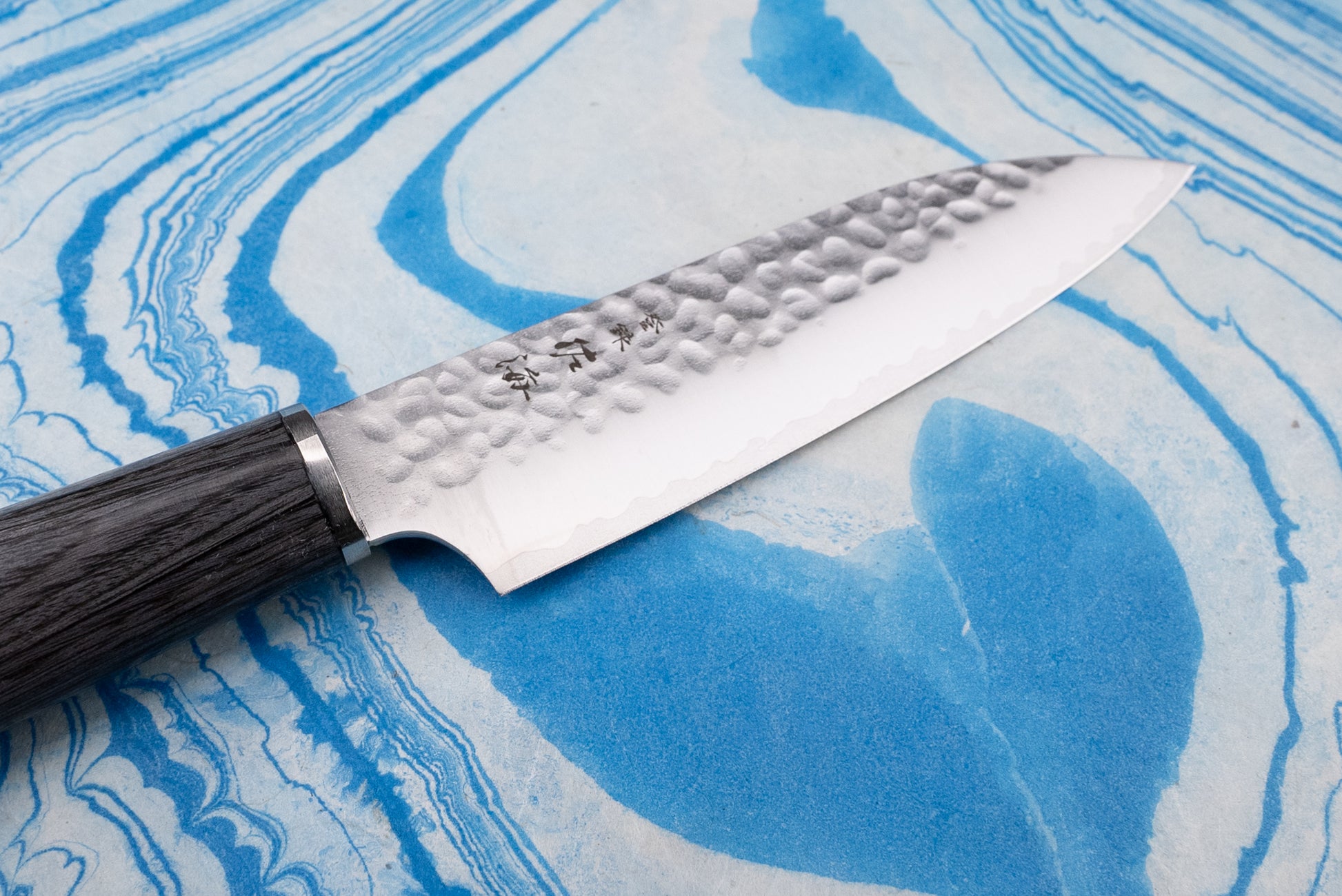 Touroku Sakai Gekka Petty 120mm: A Japanese-style gyuto with a hammered VG10 stainless steel blade and dark wooden handle, elegantly displayed on a blue-and-white marbled cutting surface.