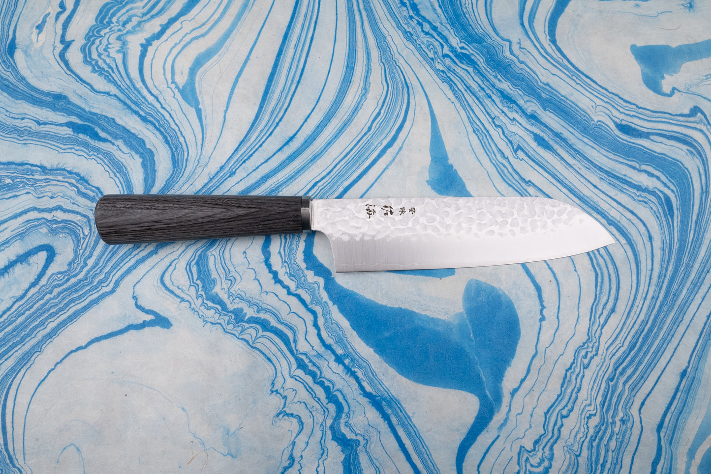 Touroku Sakai Gekka Santoku 170mm features a hammered Damascus-patterned blade with etched Japanese characters and a dark wooden handle, resting elegantly on a marbled surface.