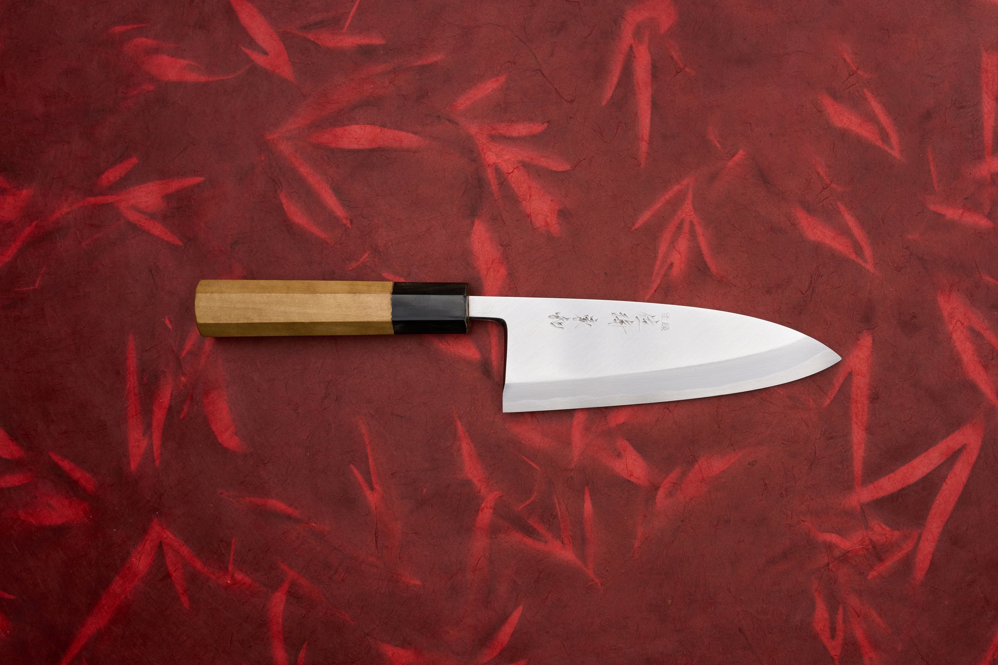 Touroku Sakai Tohwa Shirogami Deba 150mm: A Japanese santoku-style knife featuring a straight-edge stainless steel blade with an octagonal wooden handle, set against a red textured background.