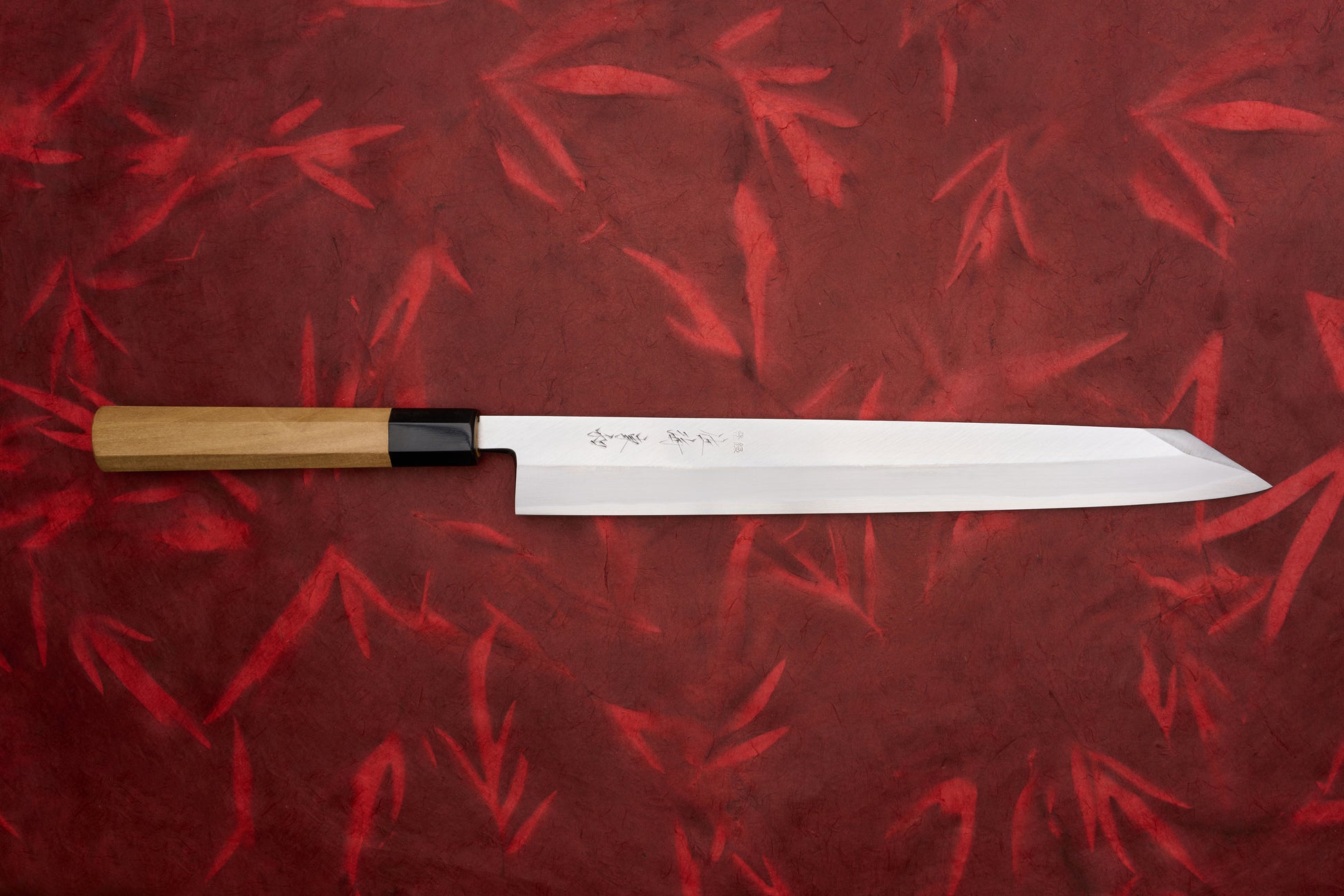 Touroku Sakai Tohwa Shirogami Kiritsuke Yanagiba 300mm, a polished Japanese slicing knife with an etched blade and octagonal wooden handle, showcased on a red textured backdrop.