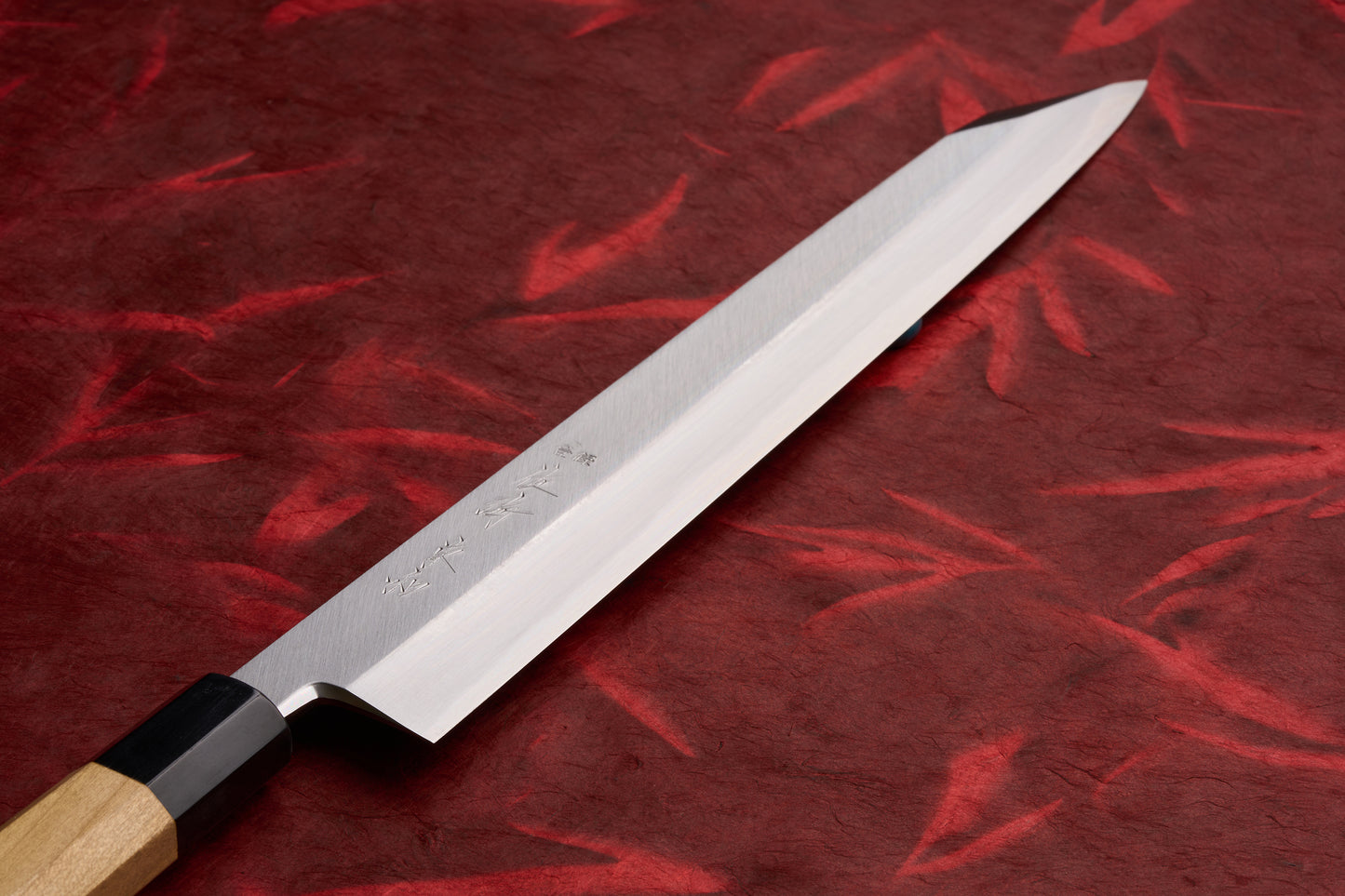 Touroku Sakai Tohwa Shirogami Kiritsuke Yanagiba 300mm knife with a high-polish, single-bevel blade featuring engraved kanji, displayed on a crumpled red background, showcasing Japanese craftsmanship.