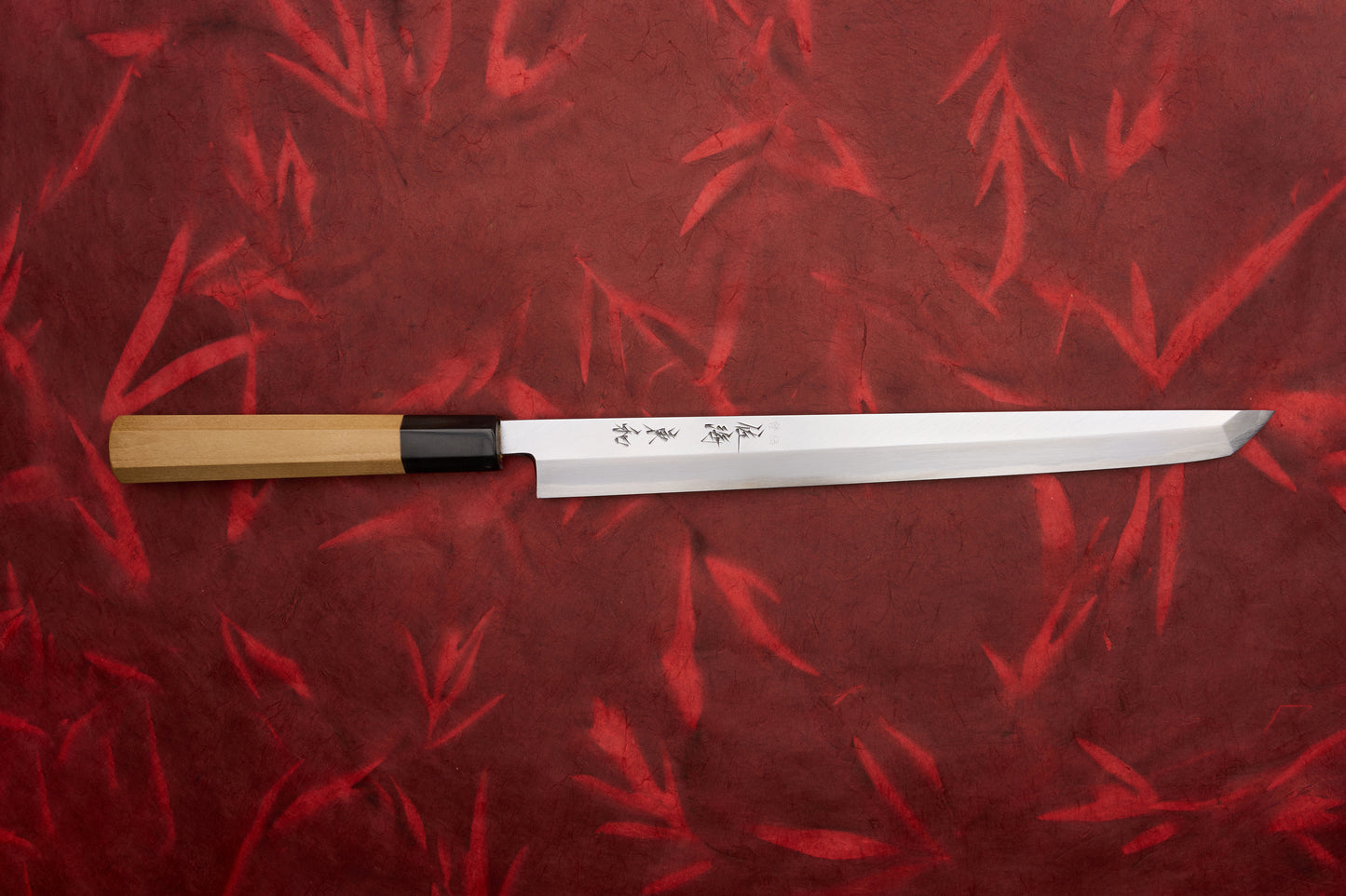 Touroku Sakai Tohwa Shirogami Sakimaru Takohiki 270mm displayed on a red, bamboo-patterned background, showcasing its slender, single-bevel polished steel blade with kanji etchings and an octagonal wooden handle.