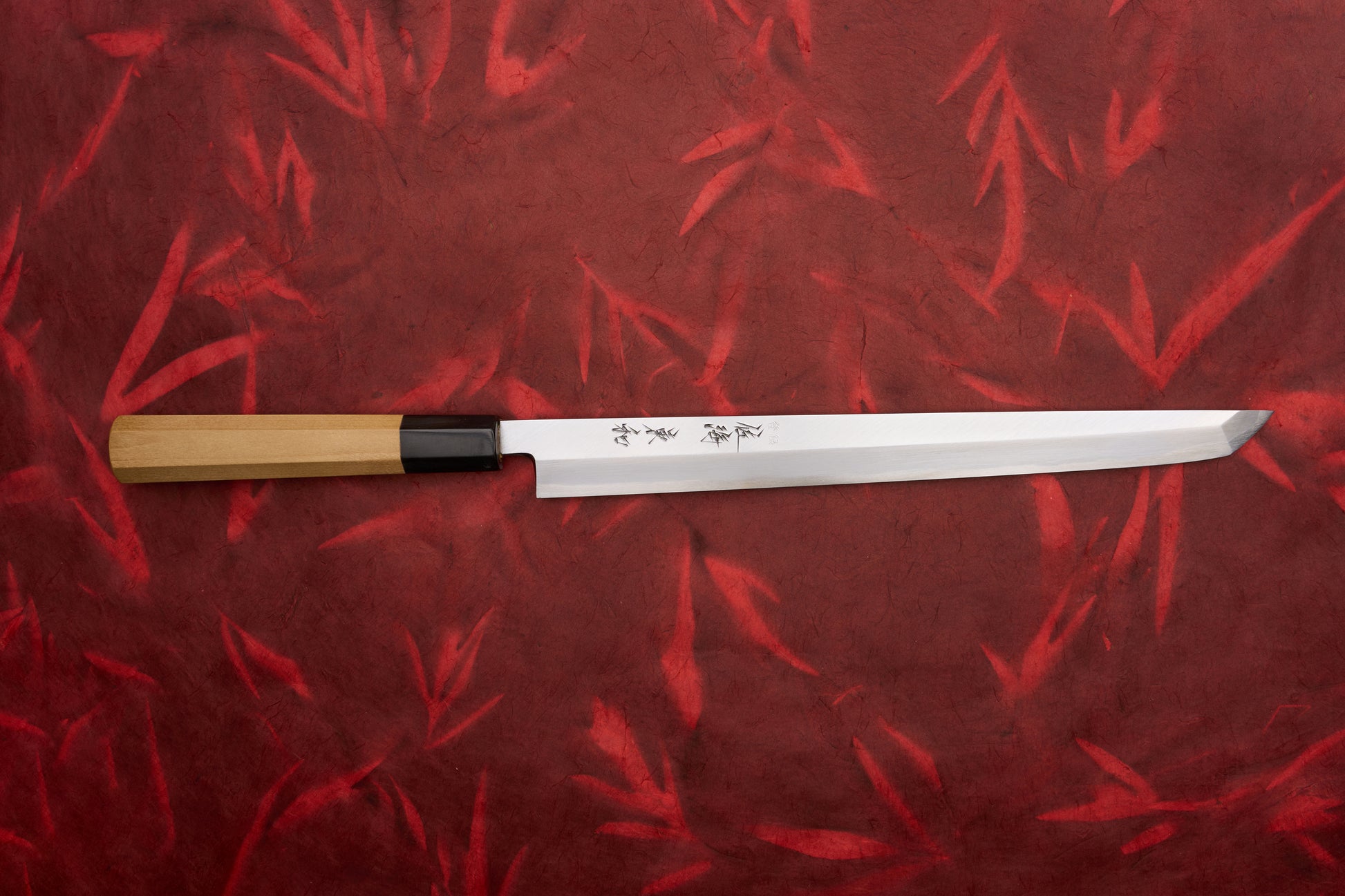 Touroku Sakai Tohwa Shirogami Sakimaru Takohiki 270mm displayed on a red, bamboo-patterned background, showcasing its slender, single-bevel polished steel blade with kanji etchings and an octagonal wooden handle.