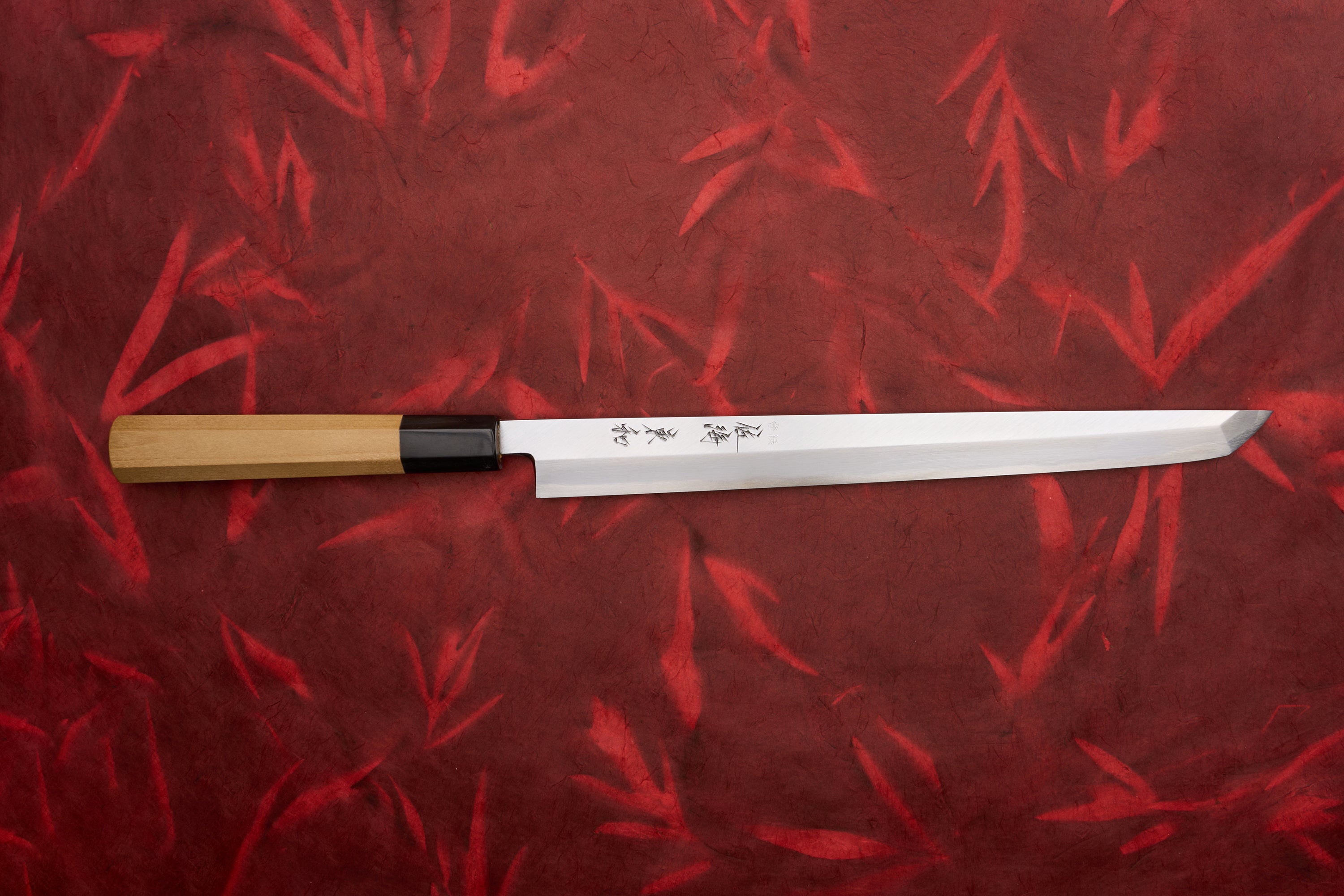 Touroku Sakai Tohwa Shirogami Sakimaru Takohiki 270mm displayed on a red, bamboo-patterned background, showcasing its slender, single-bevel polished steel blade with kanji etchings and an octagonal wooden handle.