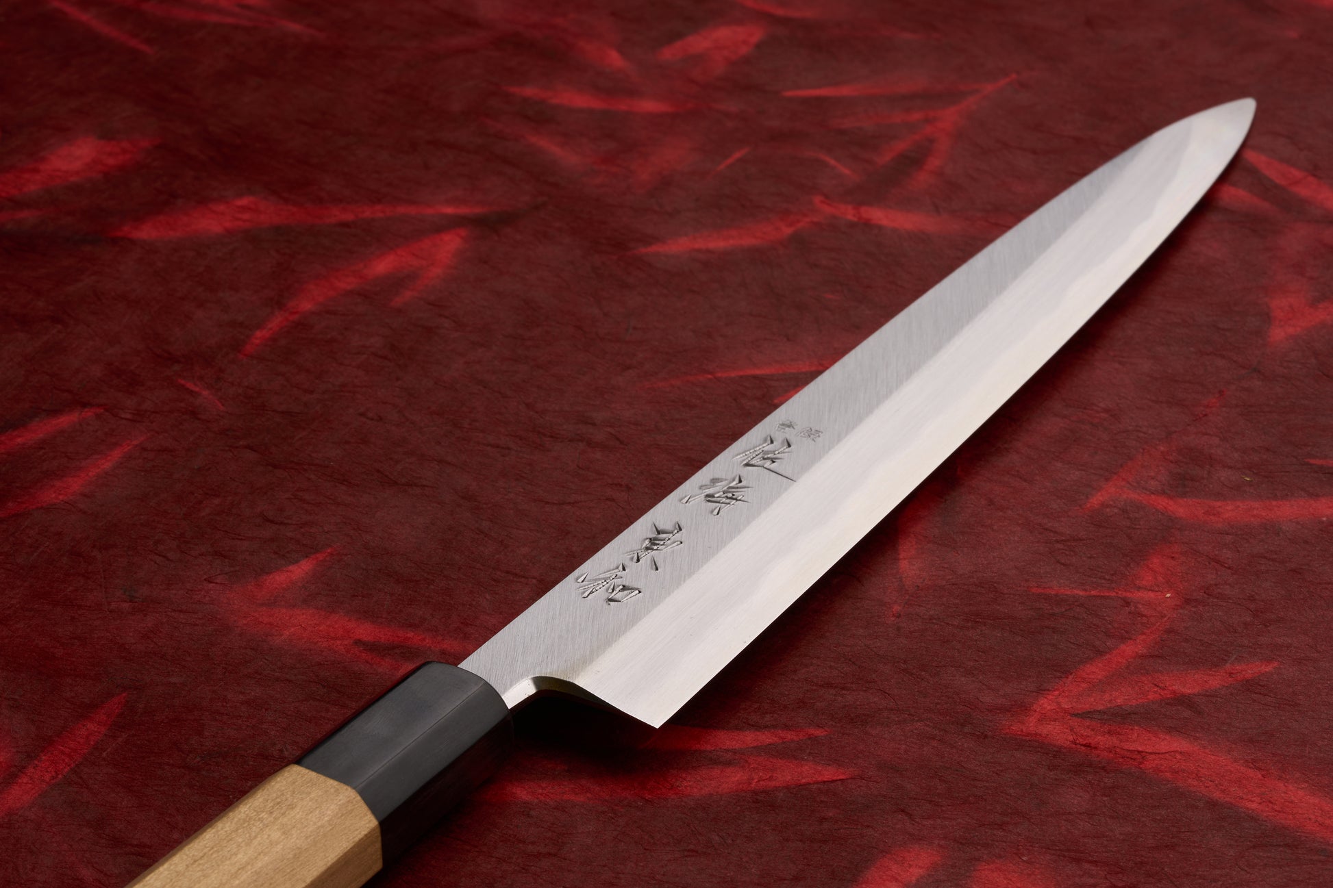 Touroku Sakai Tohwa Shirogami Yanagiba 240mm, a single-bevel sashimi knife with engraved kanji on its blade, features an octagonal wooden handle, displayed on a textured red background.