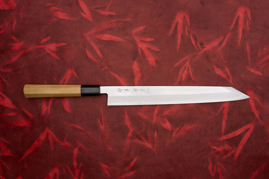 Touroku Sakai Tohwa Shirogami Kiritsuke Yanagiba 270mm knife with a sharp, single-edged blade featuring Japanese engravings, and a traditional wooden handle, displayed on a red bamboo-patterned background.