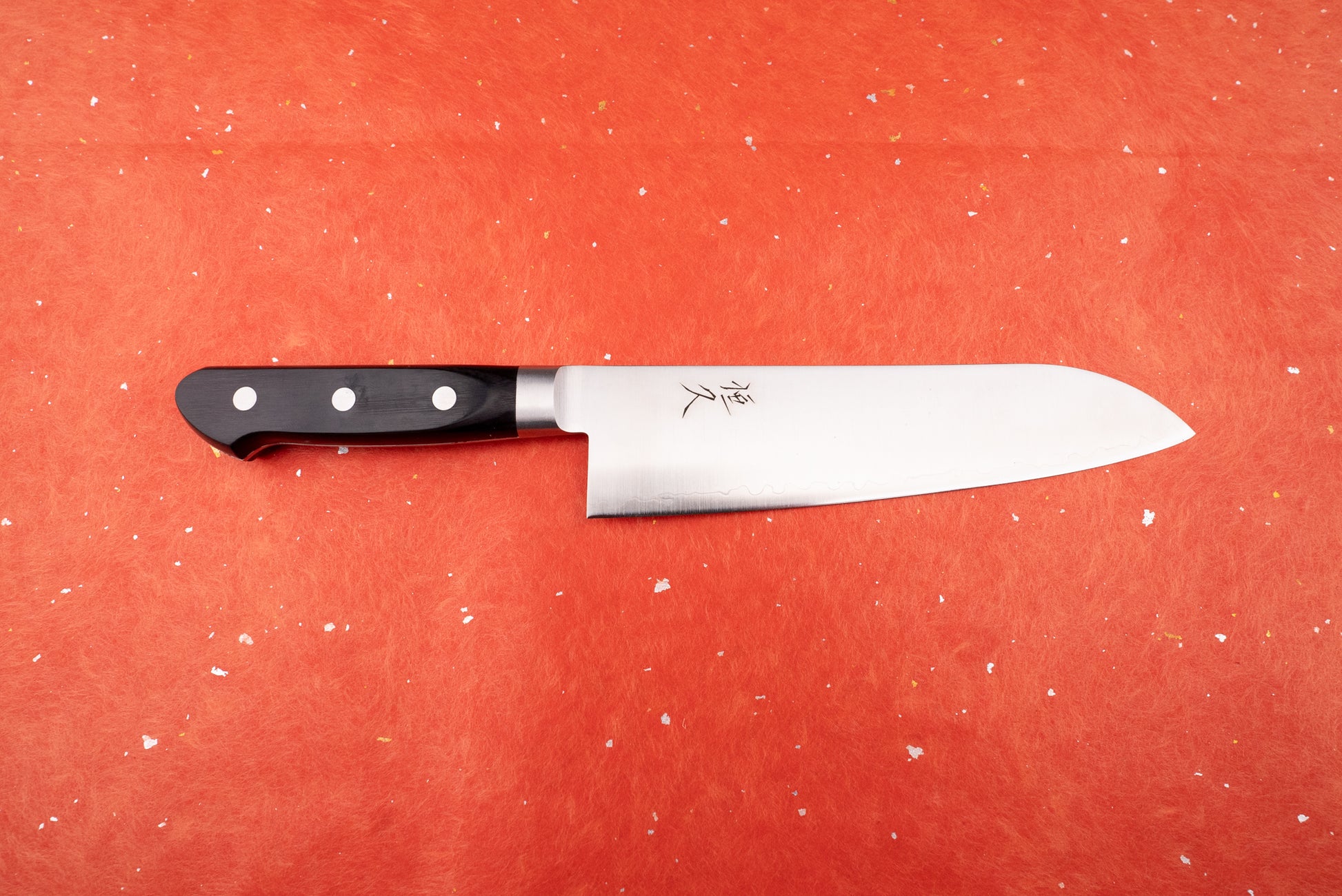 Tsunehisa 440C Sanjuni 参拾弐 Western Santoku 180mm T-03205: A Japanese-style chef's knife with a sleek steel blade, wavy temper line, and black triple-riveted handle, featured against a textured backdrop.