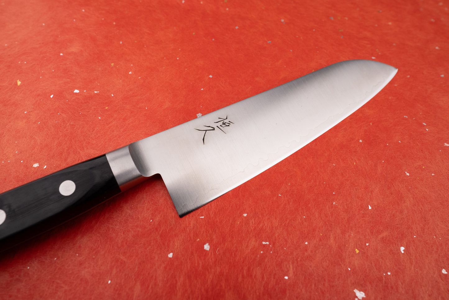 Tsunehisa 440C Sanjuni 参拾弐 Western Santoku 180mm T-03205 knife on a textured red cutting mat, featuring a hamon-style pattern, kanji engravings, and a polished black handle with visible rivets.