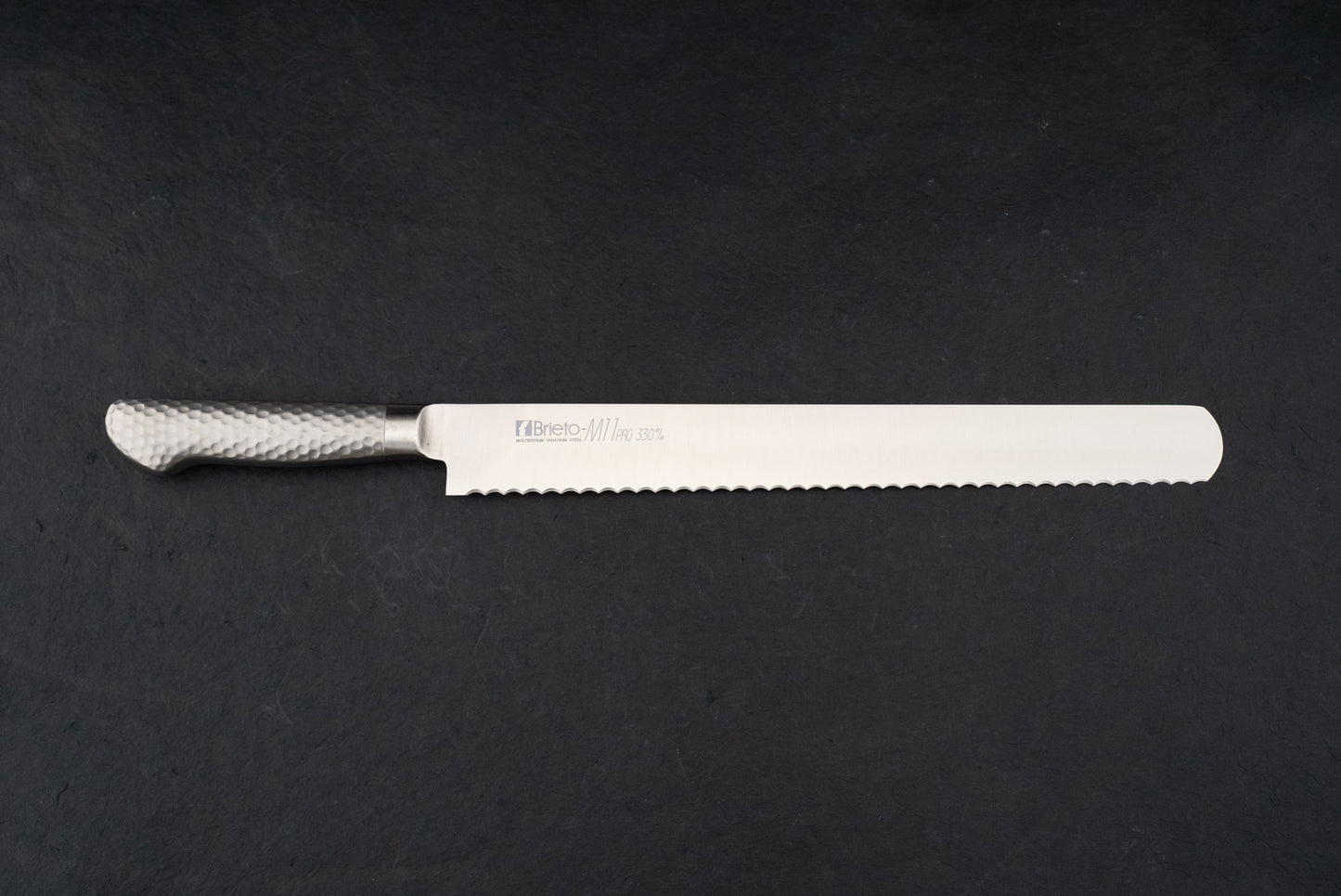 Brieto Pro Bread Knife 330mm M1150, featuring a long, serrated stainless-steel blade and a hammered-metal handle, displayed flat on a dark surface, epitomizing the precision and craftsmanship of Japanese kitchen knives.