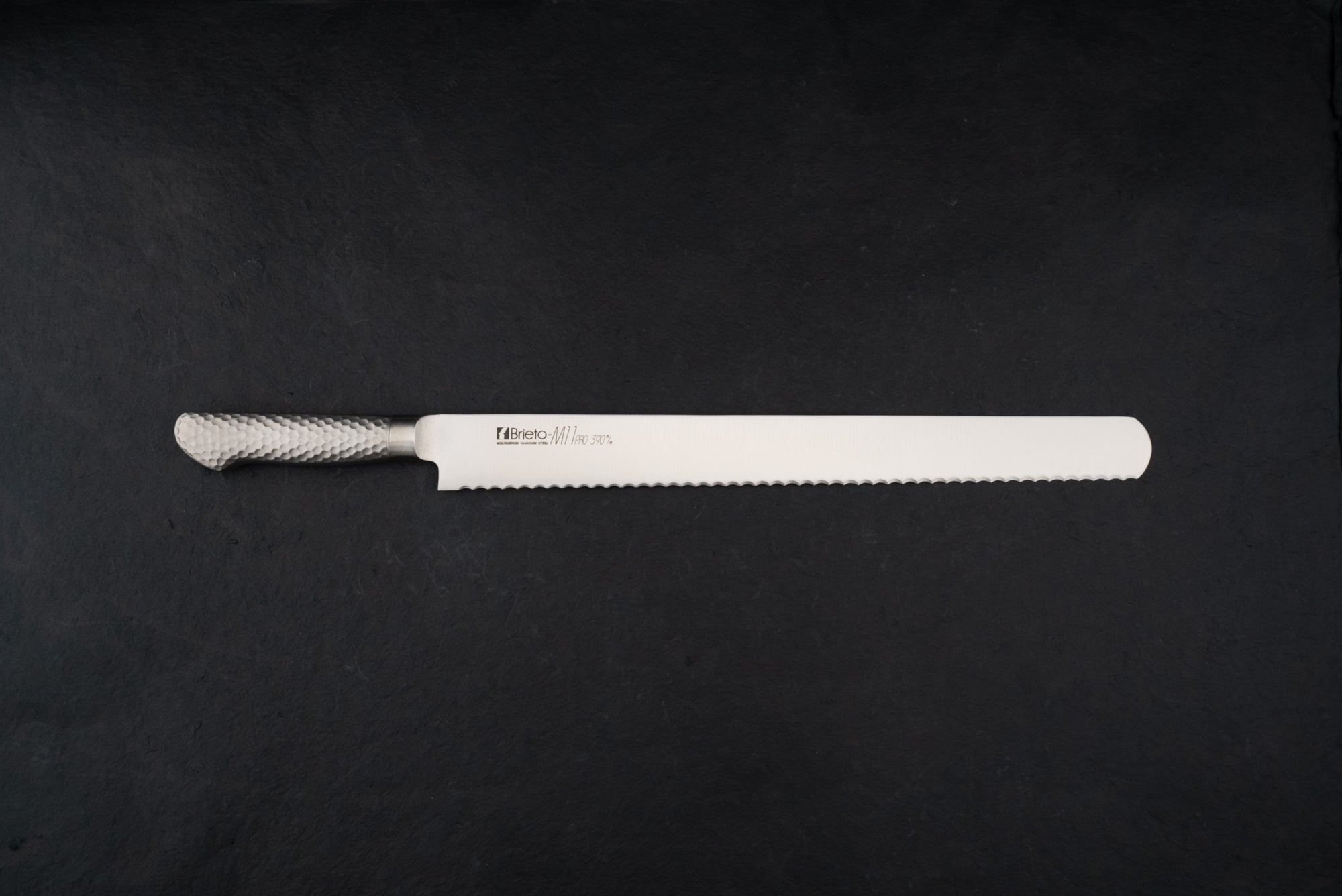 Brieto Pro Bread Knife 390mm M1148 with a long, thin serrated blade and hammered-metal handle, expertly designed for slicing bread without damaging its texture, displayed on a dark background.