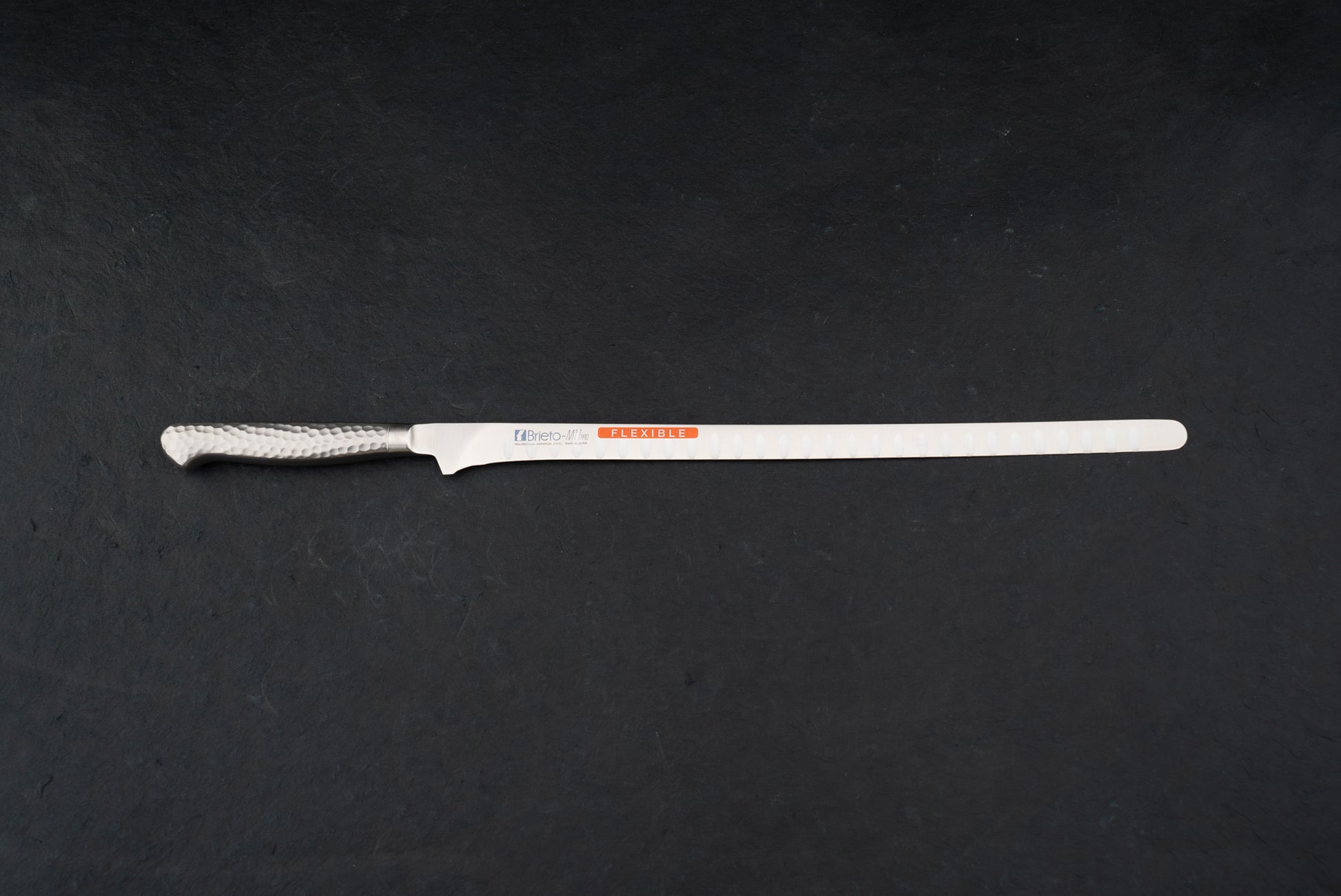 Brieto Pro Flexible Salmon Slicer with Dimples 320mm M116, featuring a long, narrow, flexible blade and hammered-finish metal handle, ideal for precise slicing of meats and fish, displayed on a dark surface.