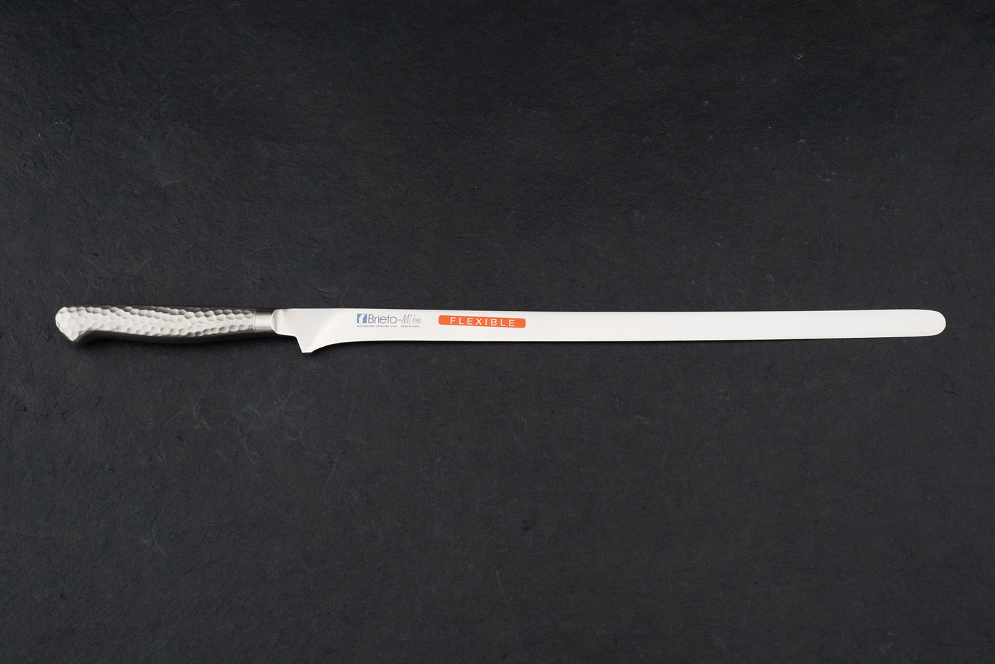Brieto Pro Salmon Slicer 320mm M115 on a countertop, featuring a slim stainless-steel blade and a hammered-steel handle, ideal for precision slicing of large meat and fish cuts.