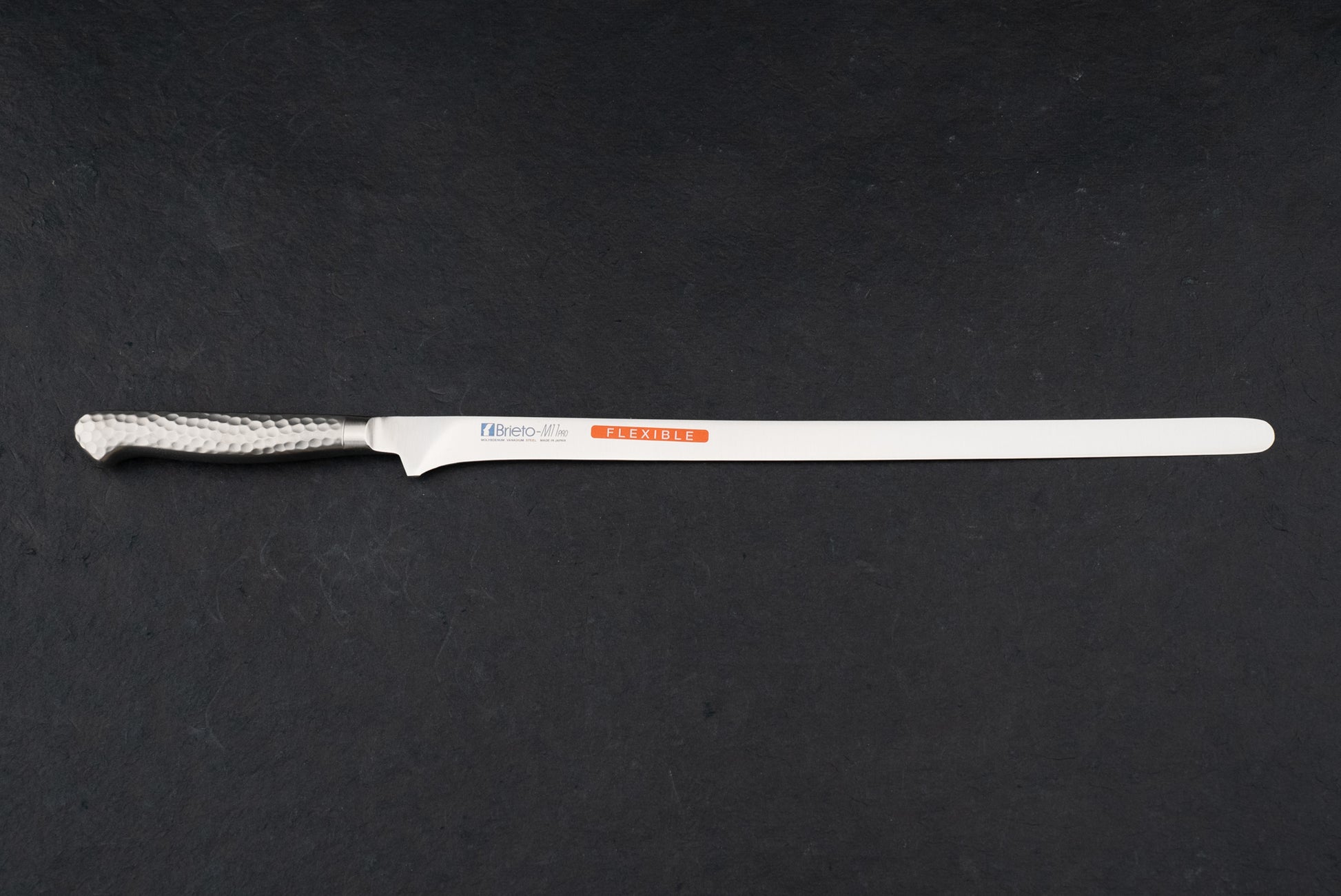 Brieto Pro Salmon Slicer 320mm M115 on a countertop, featuring a slim stainless-steel blade and a hammered-steel handle, ideal for precision slicing of large meat and fish cuts.