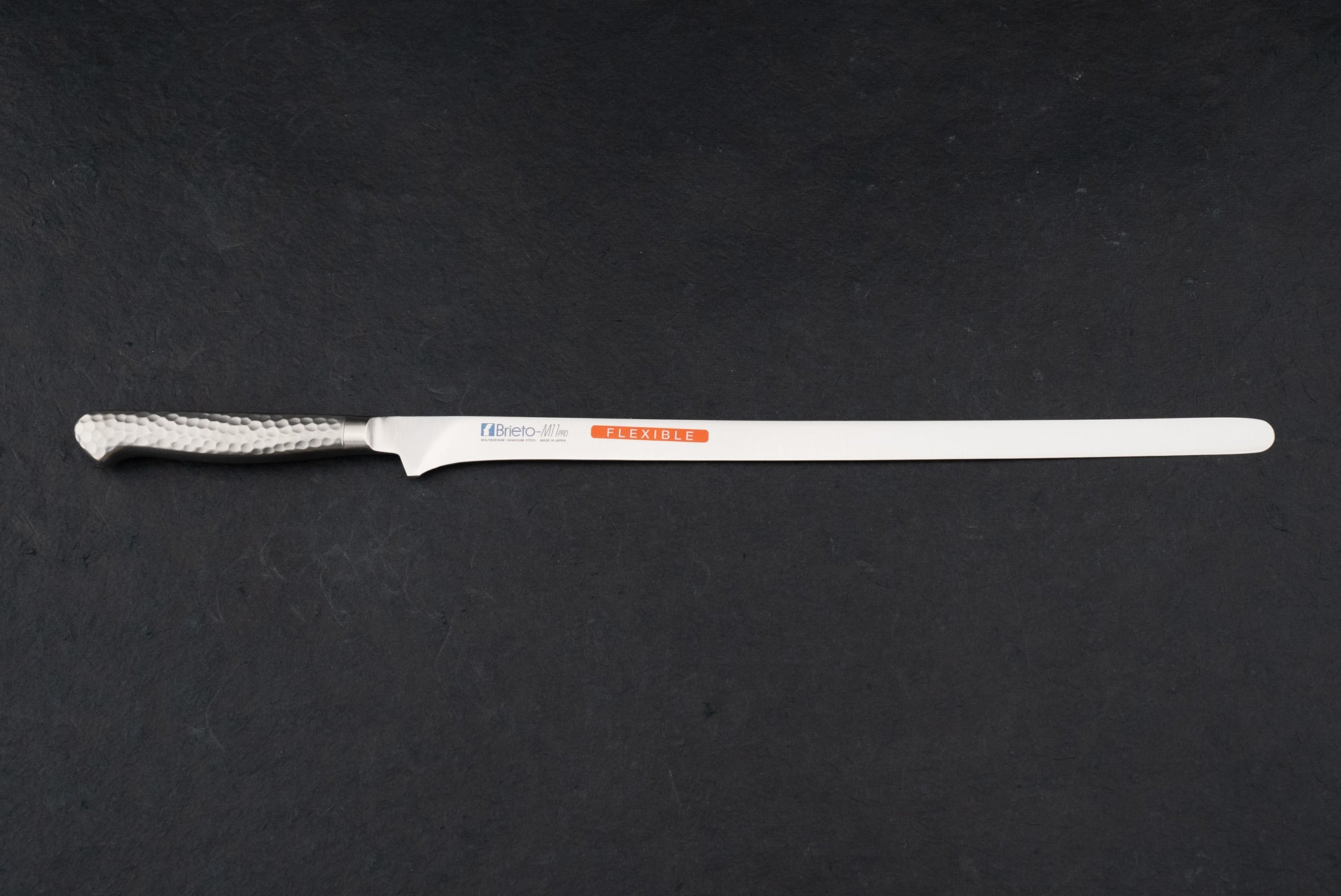 Brieto Pro Salmon Slicer 320mm M115 on a countertop, featuring a slim stainless-steel blade and a hammered-steel handle, ideal for precision slicing of large meat and fish cuts.