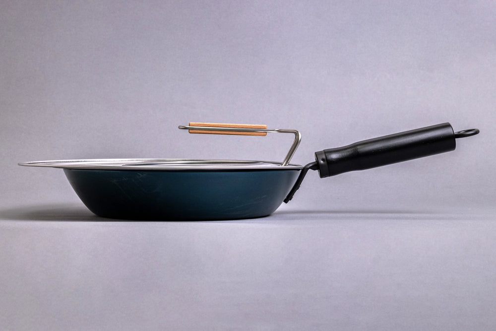 The Fujita Kinzoku Stainless Steel Universal Lid features a glass window, wooden handle, and splatter screen. It's shown with a blue non-stick frying pan and wooden-handled utensil, emphasizing Japanese craftsmanship.
