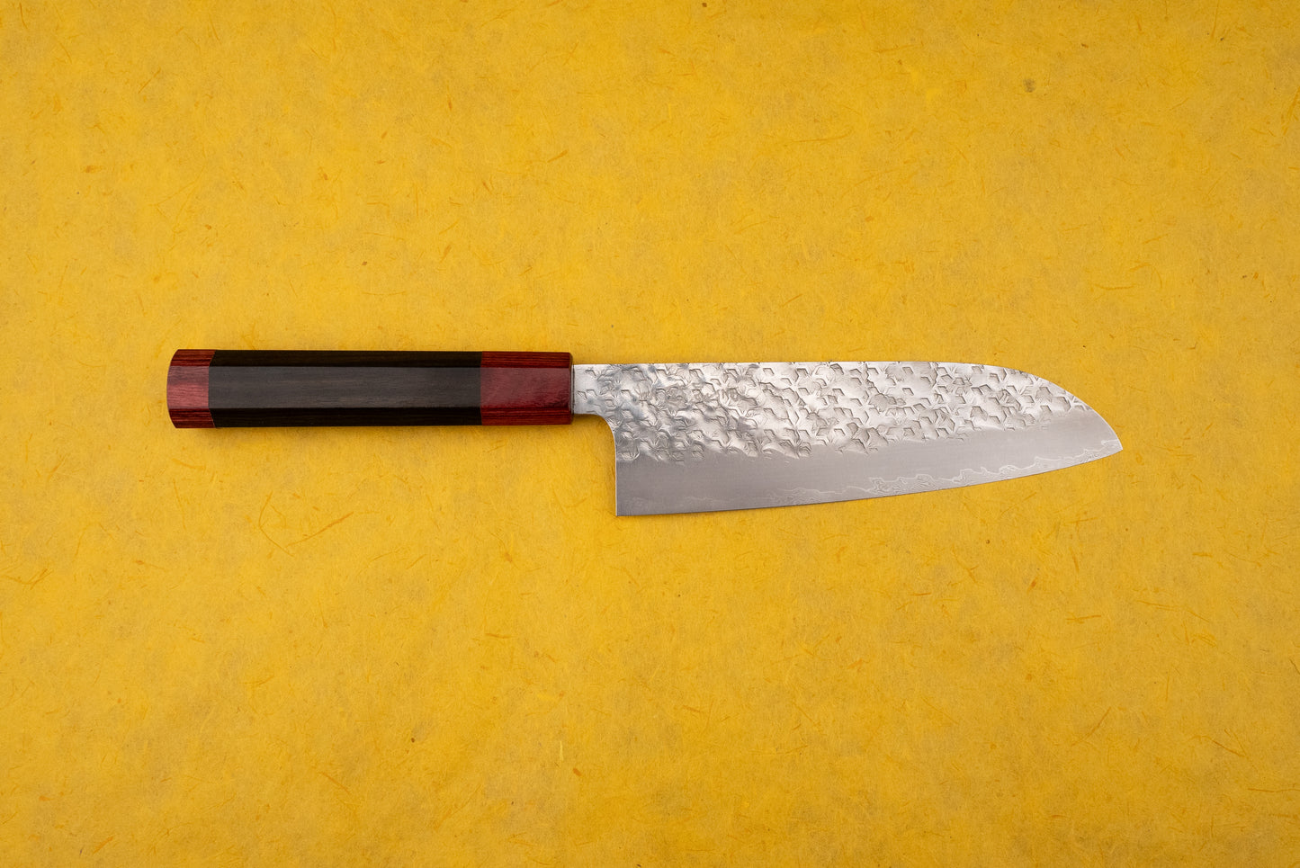 Makoto Kurosaki Coreless Kaen Tsuchime Santoku 165mm knife on yellow background, features a hammered Damascus blade and octagonal dark wood handle with red accents, epitomizing Japanese precision and craftsmanship.