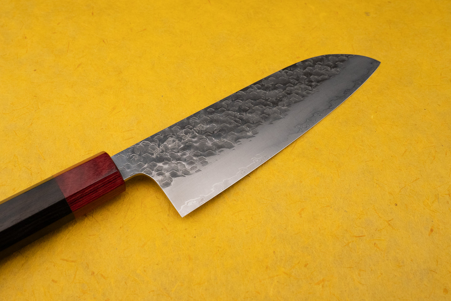 Makoto Kurosaki Coreless Kaen Tsuchime Santoku 165mm knife on a yellow surface, showcasing its layered Damascus steel blade with hammered finish and traditional octagonal wooden handle with red ferrule.