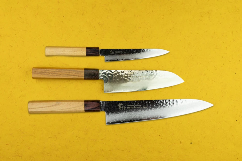Sakai Takayuki VG10 33 Layer Damascus 3 Piece Knife Set, featuring a petty, santoku, and gyuto knife with hammered steel blades and rectangular wooden handles, elegantly displayed on a bright yellow background.