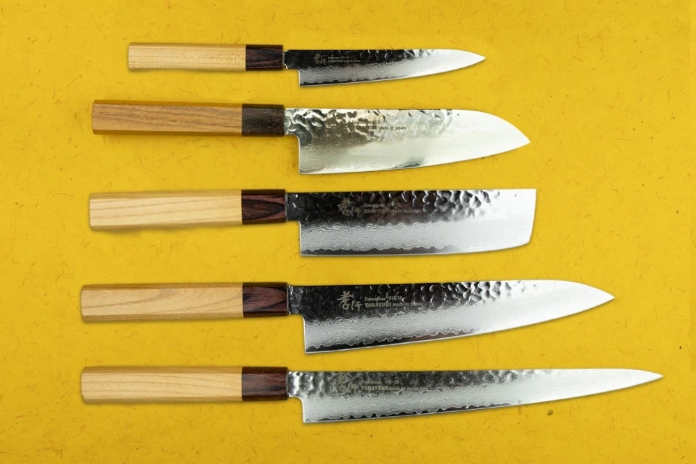 Sakai Takayuki VG10 33 Layer Damascus 5 Piece Knife Set displayed on yellow. Includes petty, santoku, nakiri, gyuto, and sujihiki knives, showcasing hammered stainless-steel blades and wooden handles, embodying Japanese craftsmanship.