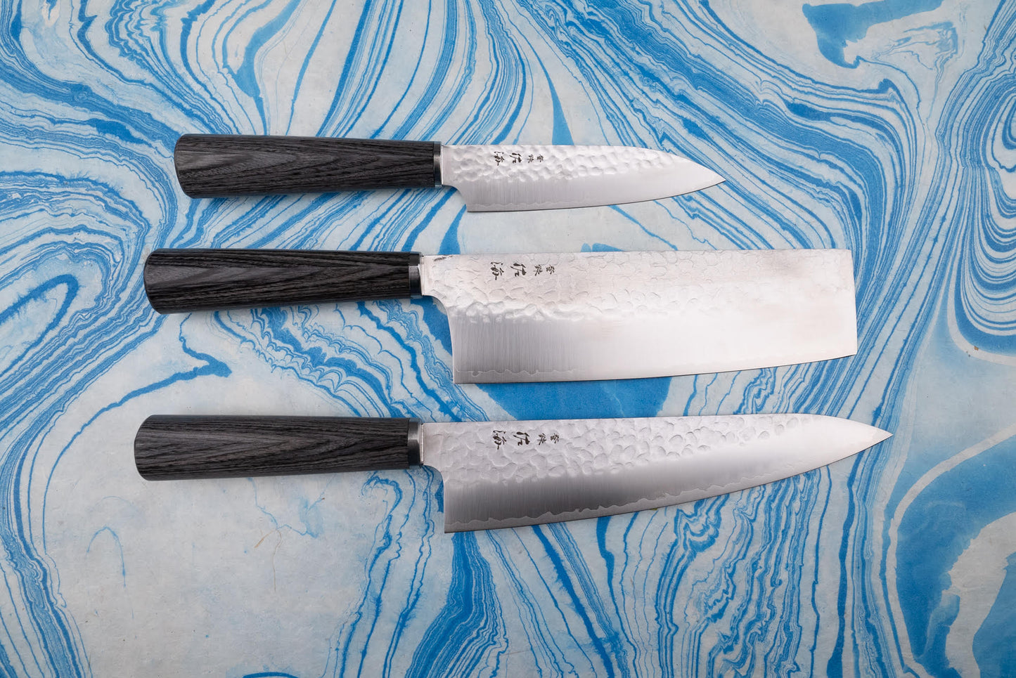 Touroku Sakai Gekka 3 Piece Knife Set on a marbled surface, featuring a hand-hammered petty knife, nakiri, and gyuto with dark wood handles, showcasing precision craftsmanship and exceptional balance.